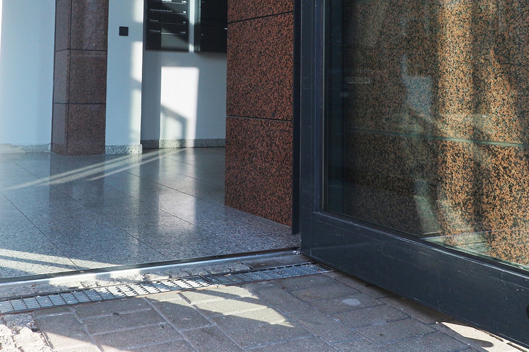 The entrance door made of glass has an anthracite frame and is open; in the foreground you can see the paving stones of the street and a threshold to the entrance area, in front of which there is a water grate; the floor in the entrance area is speckled gray, the walls are white with tile elements speckled black-dark red; in the background you can see anthracite mailboxes