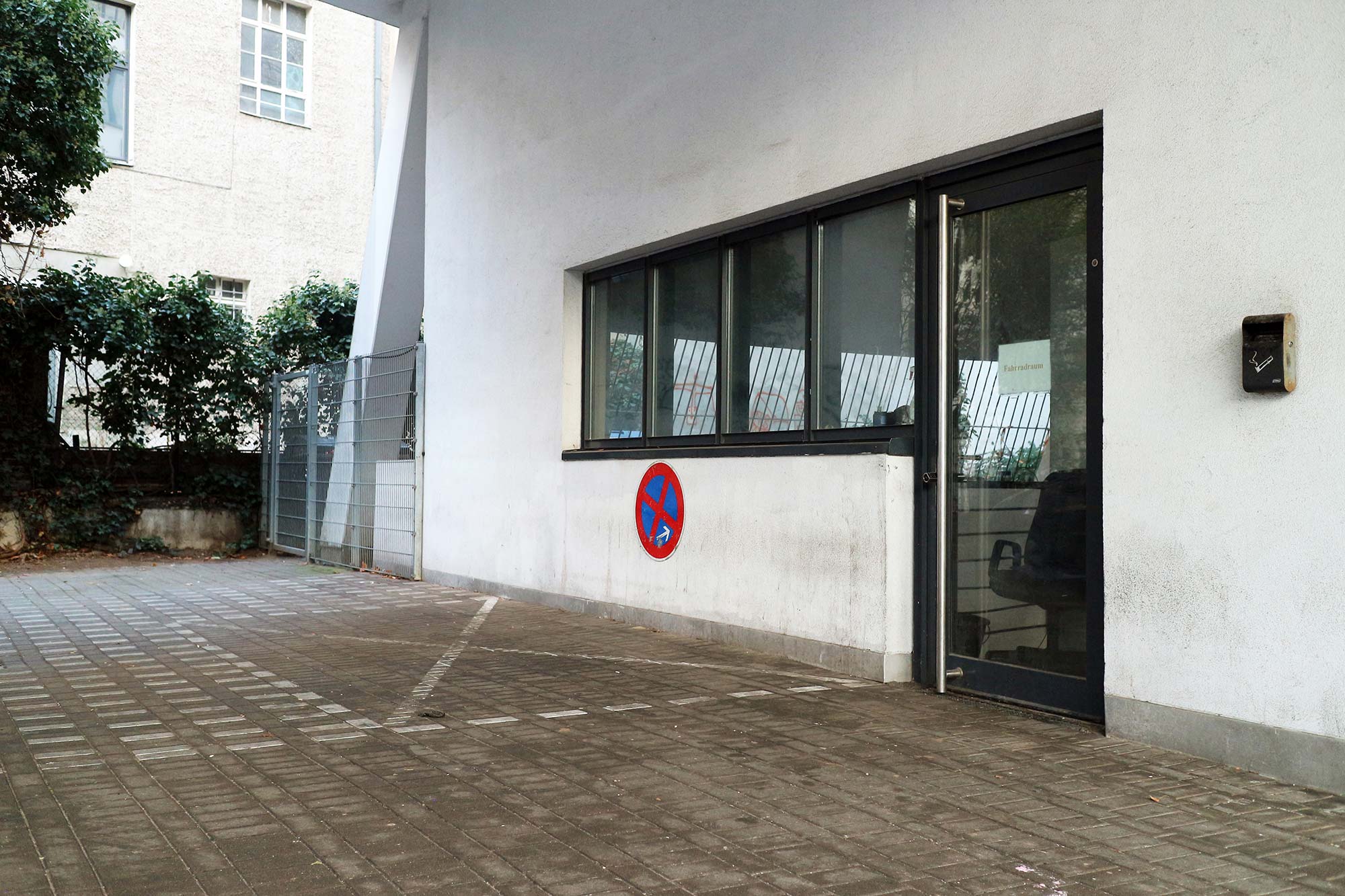 A covered area with cobblestones can be seen with a marked parking space in the center; the building to the right has a white wall and a window front with a glass door; below the windows there is a no stopping sign with an arrow pointing to the right; in the background a bush can be seen behind which there is another office building