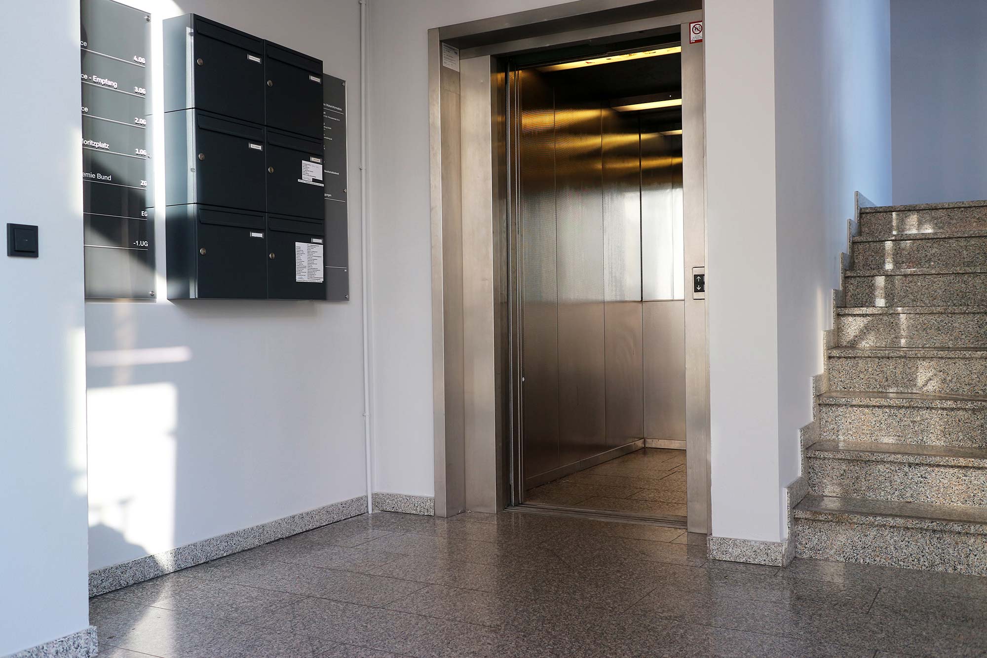 The entrance area of the office building has white walls and a mottled gray floor; anthracite mailboxes hang on the wall to the left; to the right, steps go up to the second floor; in the middle the elevator, whose door is open, is located; the elevator is silver