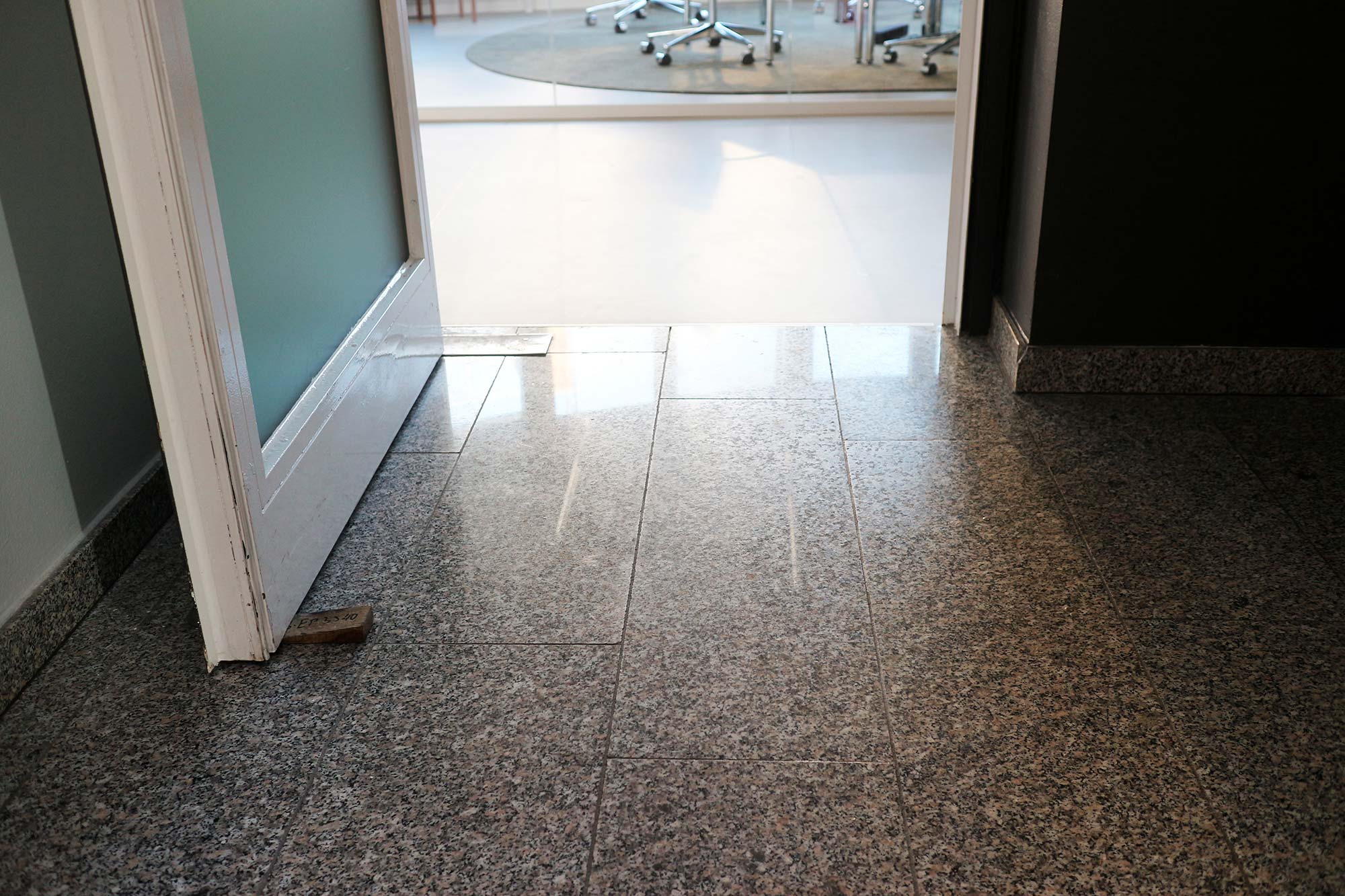 A white entrance door with frosted glass in the DigitalService office, held open by a wooden wedge; the wall on the left is white, the wall straight out next to the door on the right is anthracite; the floor is tiled with black and white speckled tiles; legs from chairs from a meeting room can be seen in the background