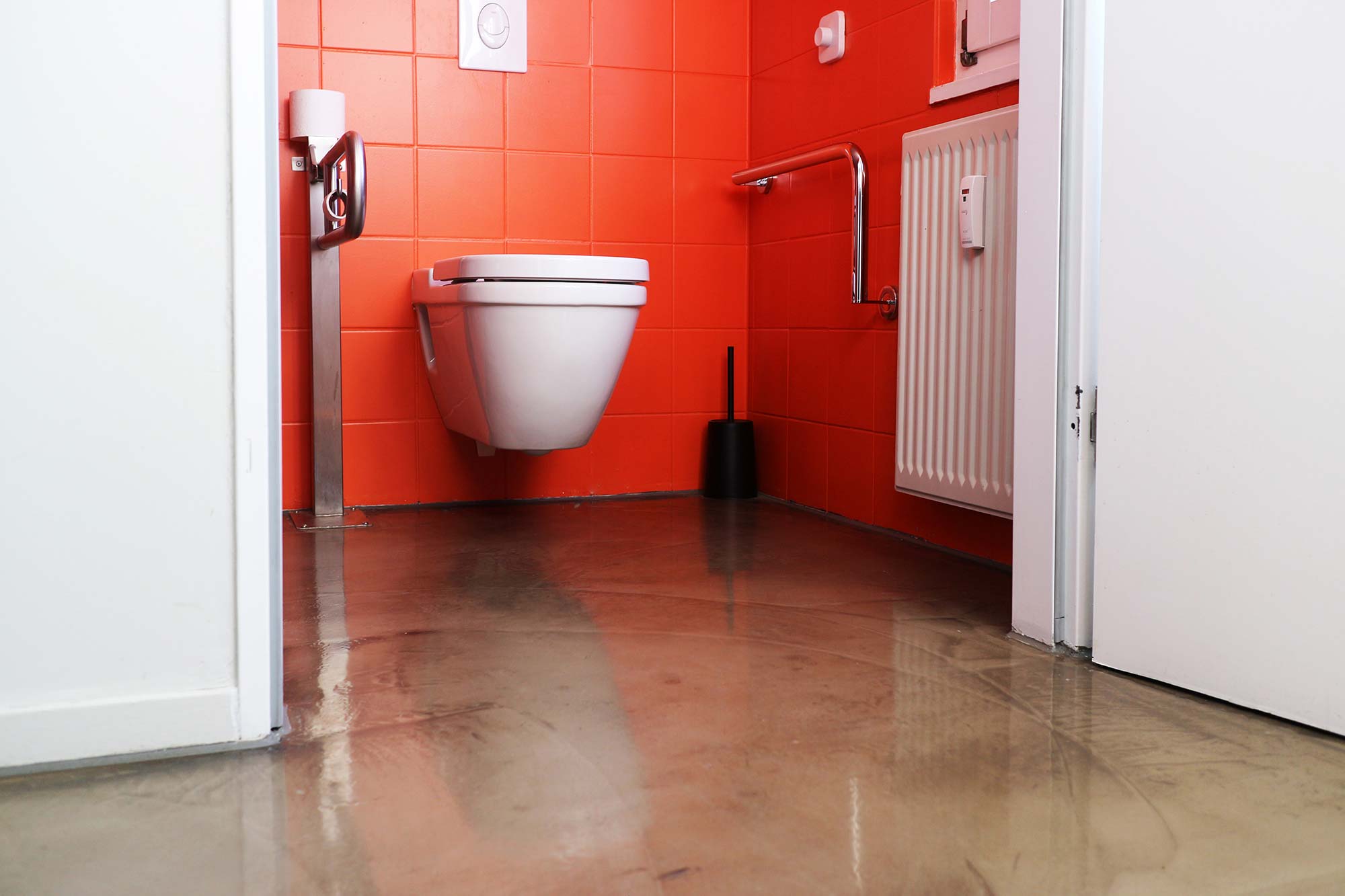 The wide white door to the accessible toilet is open and shows the white toilet next to which there are two silver safety trails; the wall is tiled in a bright orange and the floor has a shiny beige color; on the right side on the wall just behind the door is a small white radiator