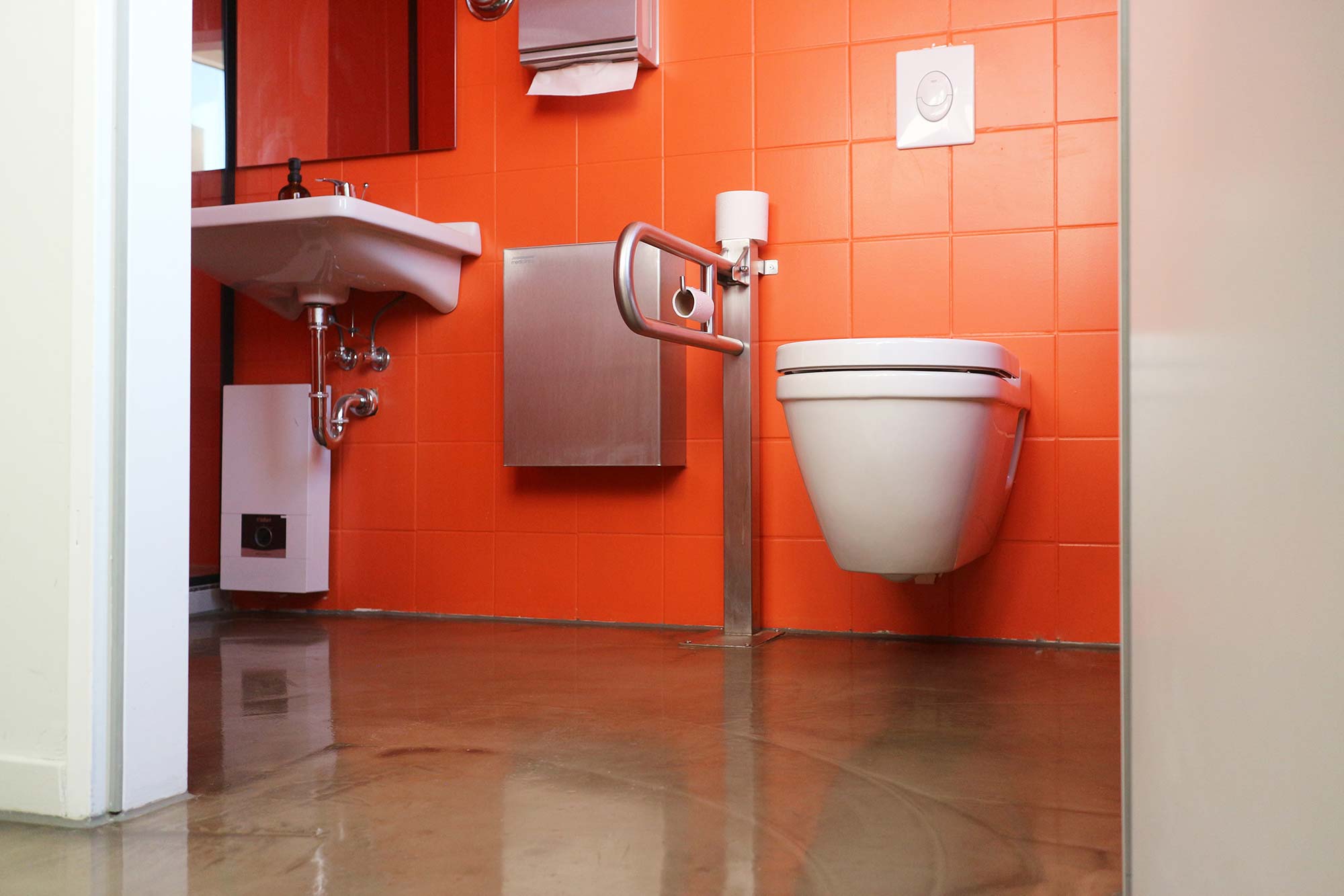 The accessible toilet is tiled with bright orange tiles; the fixtures (toilet and sink) are white, while the toilet safety rail, the wall-mounted trash can and the towel dispenser are silver; a mirror hangs above the sink