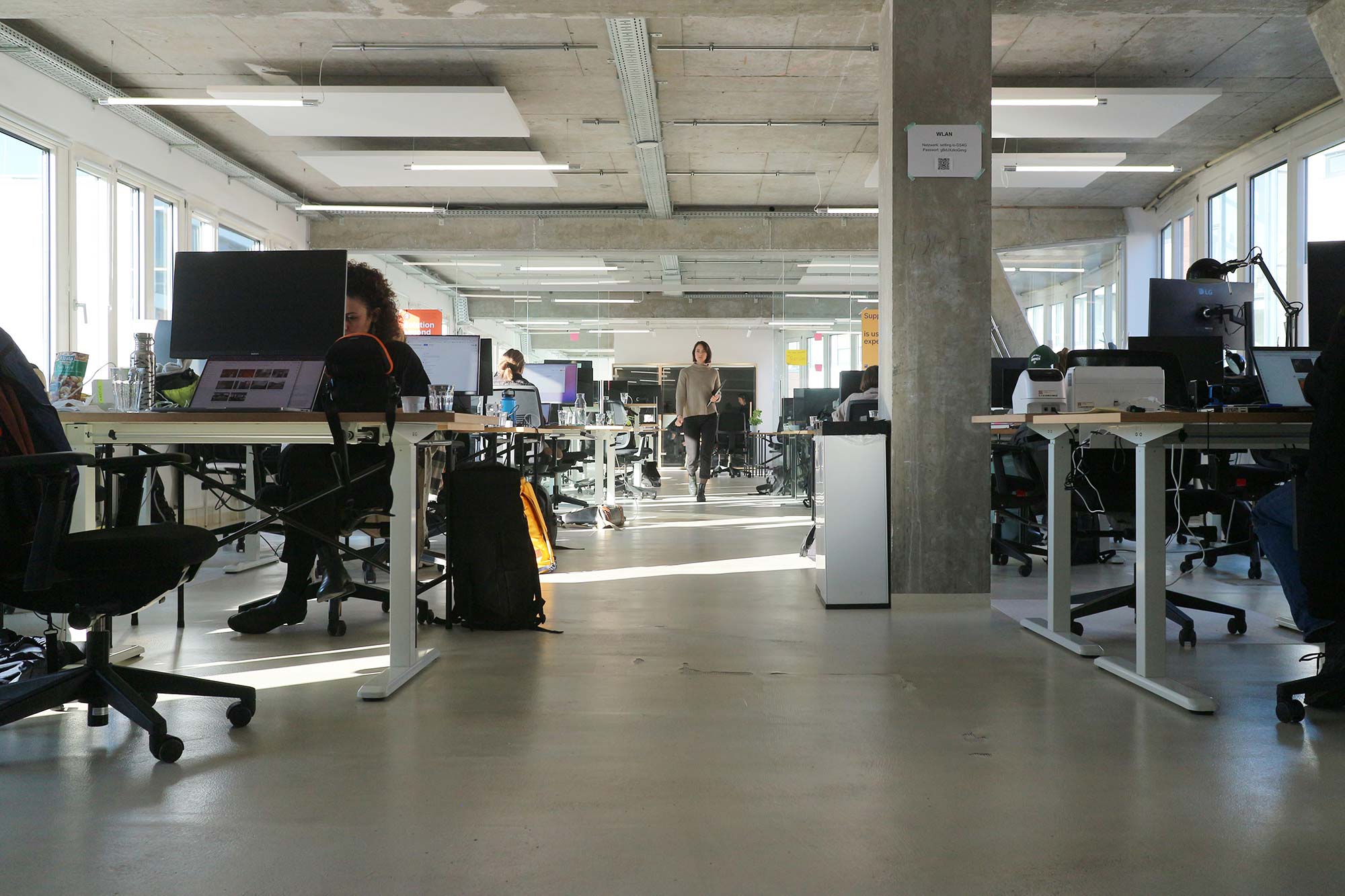 A large open office space with a hallway in the middle; large work desks with tables are on the right and left; there are screens, laptops, and other office supplies on the desks; a person walks from back to front in the hallway; the ceiling is exposed concrete, the floor is gray, and there are windows on the side walls