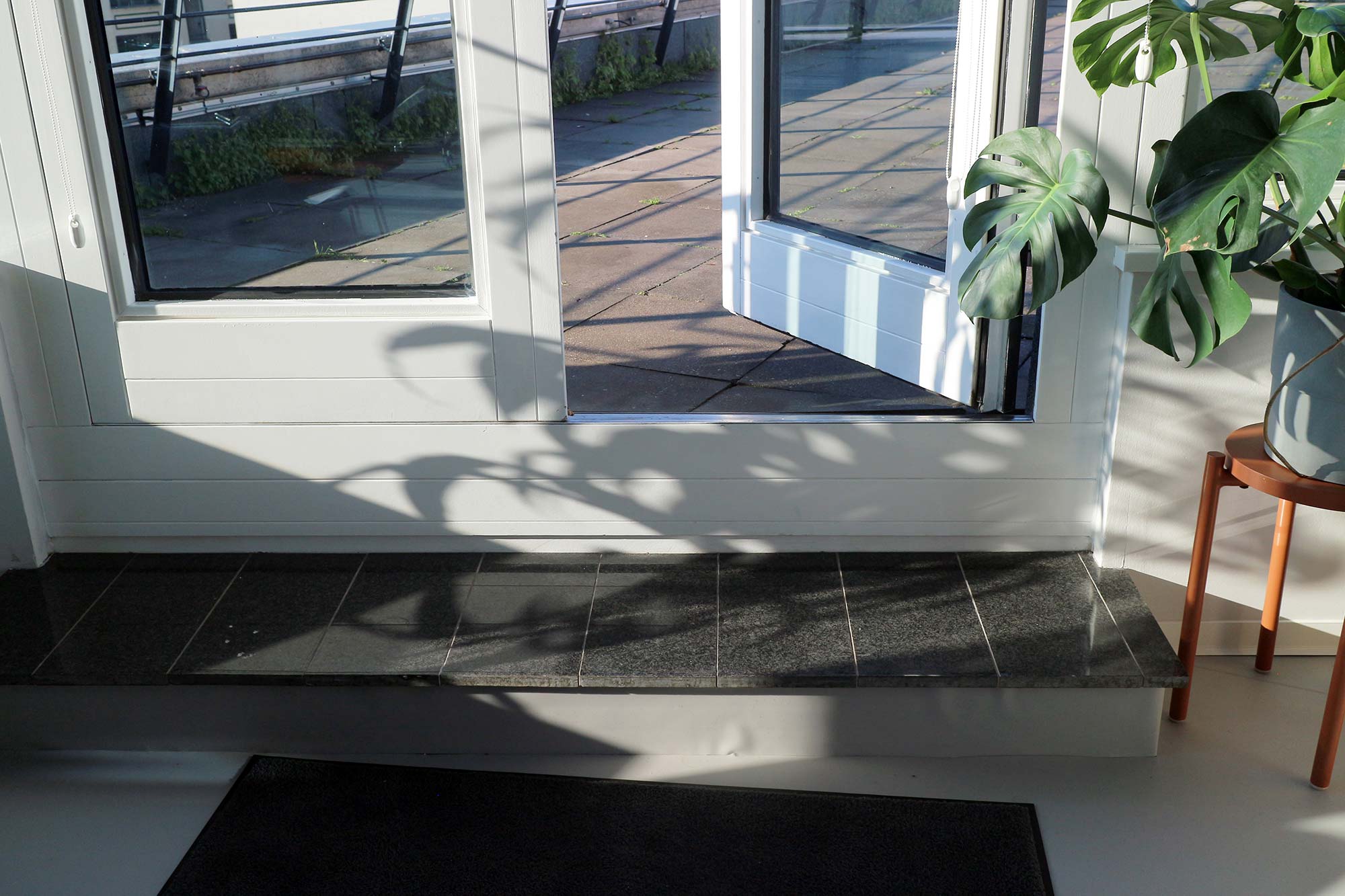 The exit to the roof terrace on the fourth floor that can be seen consists of two glass doors with a white frame, the right one is open; a doormat is inside and the step to the exit is tiled in black; to the right of the doors there is a green plant on a small wooden table