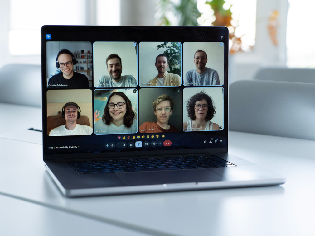 A laptop stands on a table in an office; the screen shows eight people in a video call who are part of the “Accessibility Ambassadors” working group