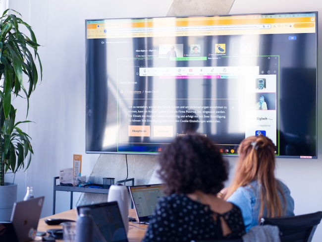 Two women sit in front of laptops in a modern office, while a large screen displays a video conference and a website. A houseplant stands to the left of the room.