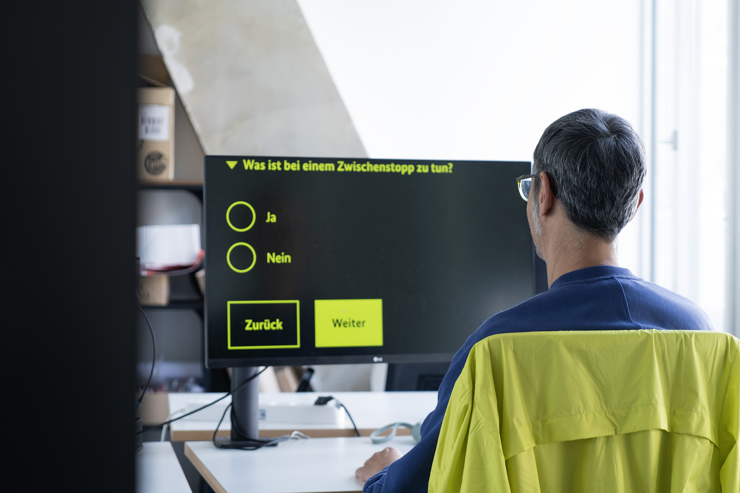 A man sits in front of a monitor on which a multiple-choice question is displayed in German in greatly enlarged form: “What should I do if I make a stopover?” with the options “Yes” and “No” as well as “Back” and “Next” buttons.