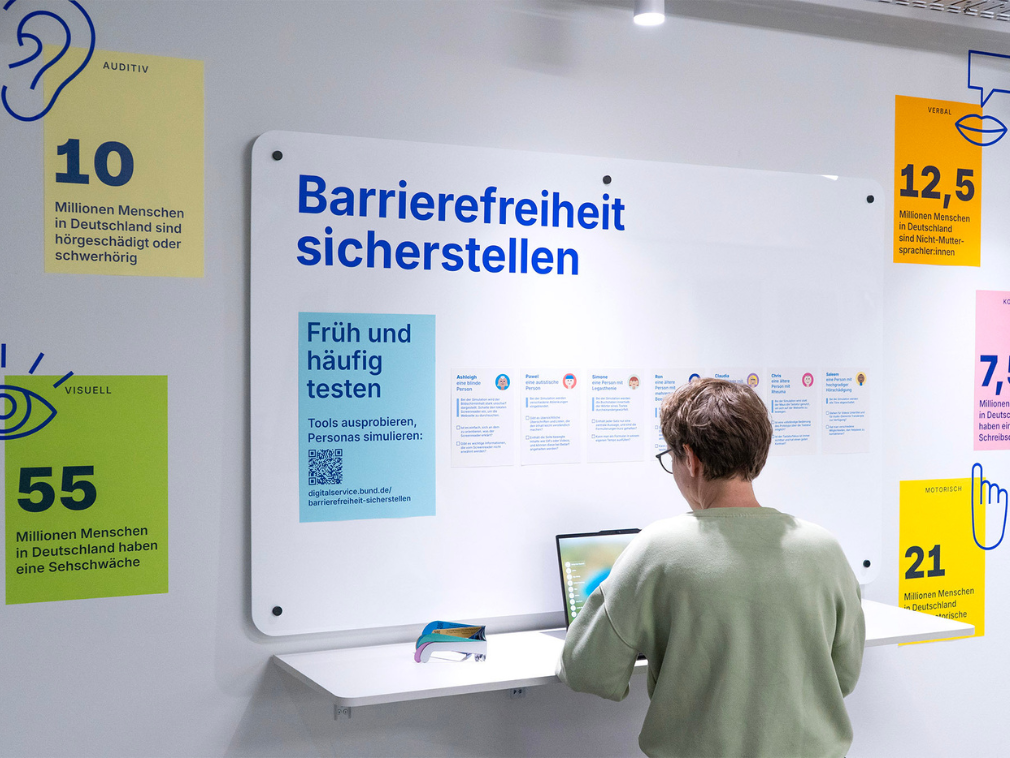A person sits at a table with a laptop in front of a large white information board with the heading “Ensuring accessibility” in the DigitalService empathy lab. The board displays information on various disabilities and statistics in Germany, such as “10 million people in Germany are hearing impaired or hard of hearing”.