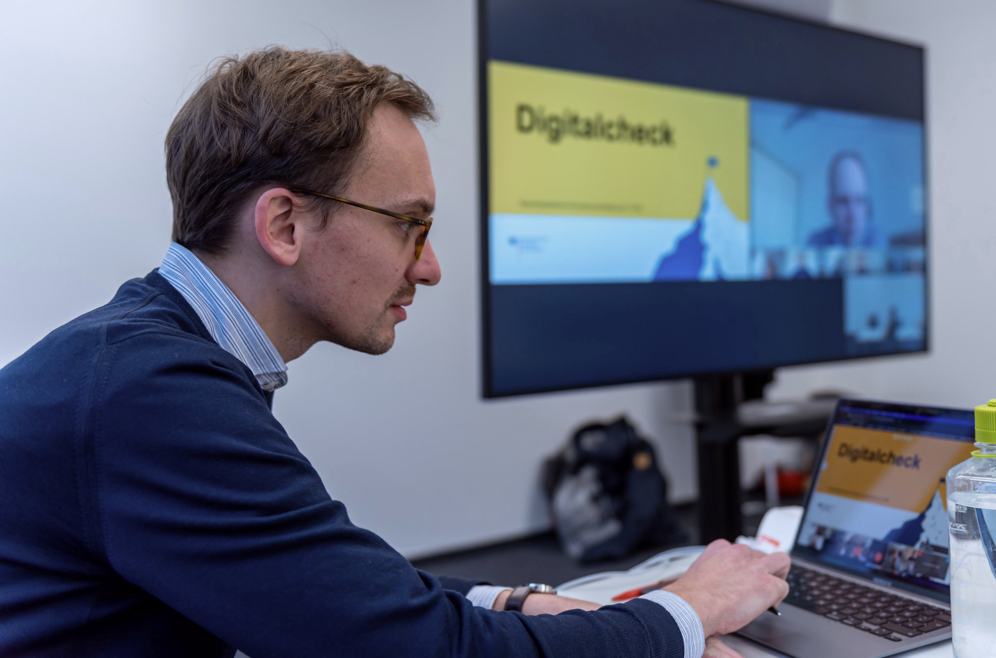 Eine Person nimmt an einem virtuellen Meeting teil. Sowohl auf ihrem Laptop-Bildschirm als auch auf einem großen Bildschirm im Hintergrund ist neben weiteren Teilnehmenden der Schriftzug „Digitalcheck“ gut zu erkennen.