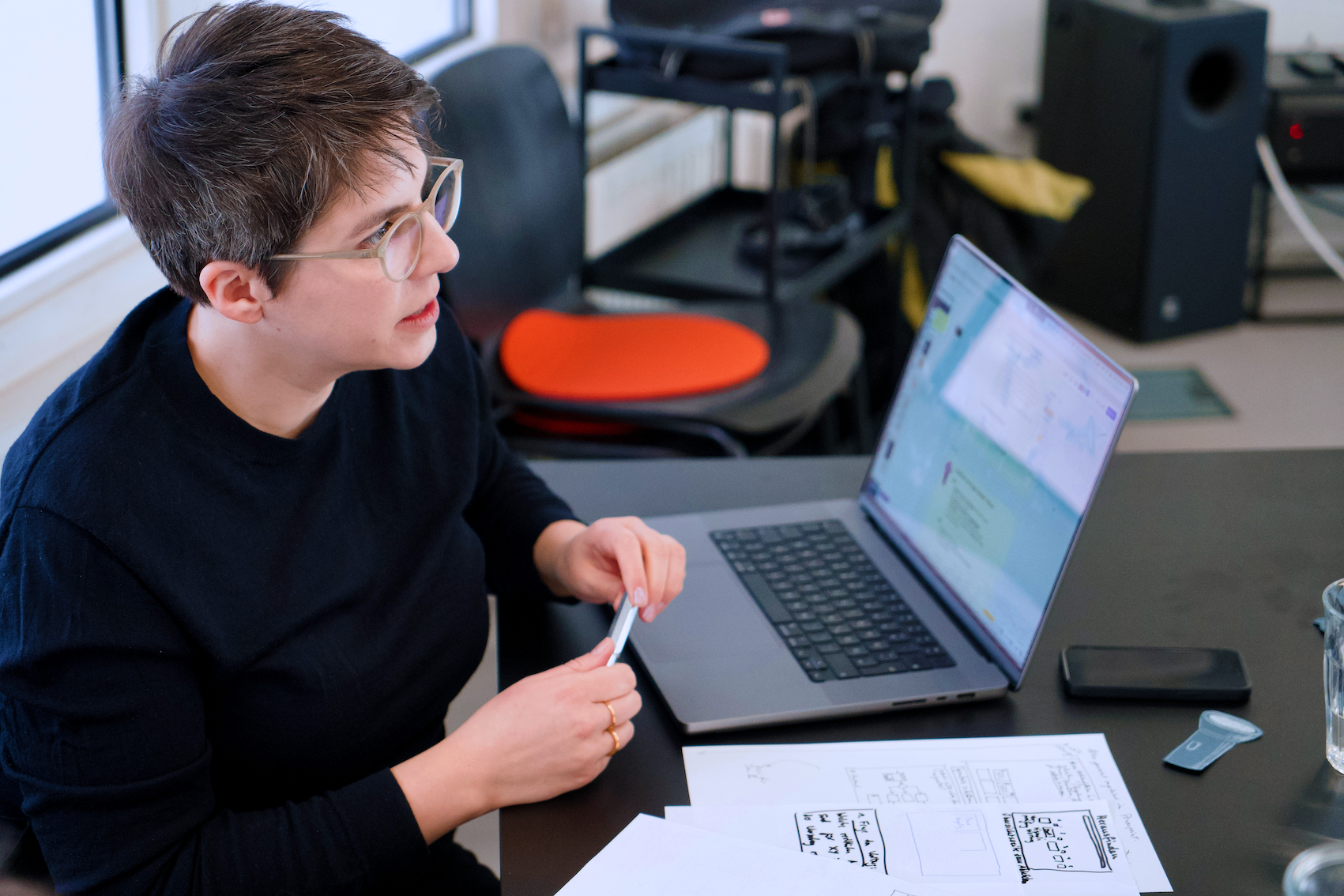 A person is sitting at a black desk in a modern office space, holding a pen and talking. There is a laptop in front of the person. A program is running on the open screen. There are notes and a cell phone on the table. Office objects can be seen in the background.