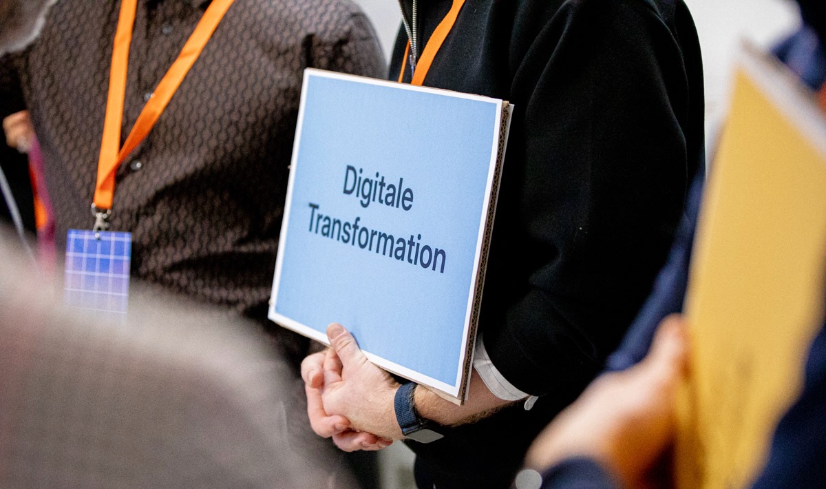 Person holding a sign with the words “Digital Transformation”. The person is wearing a black top and an orange lanyard. Other people with name badges can be seen in the background.