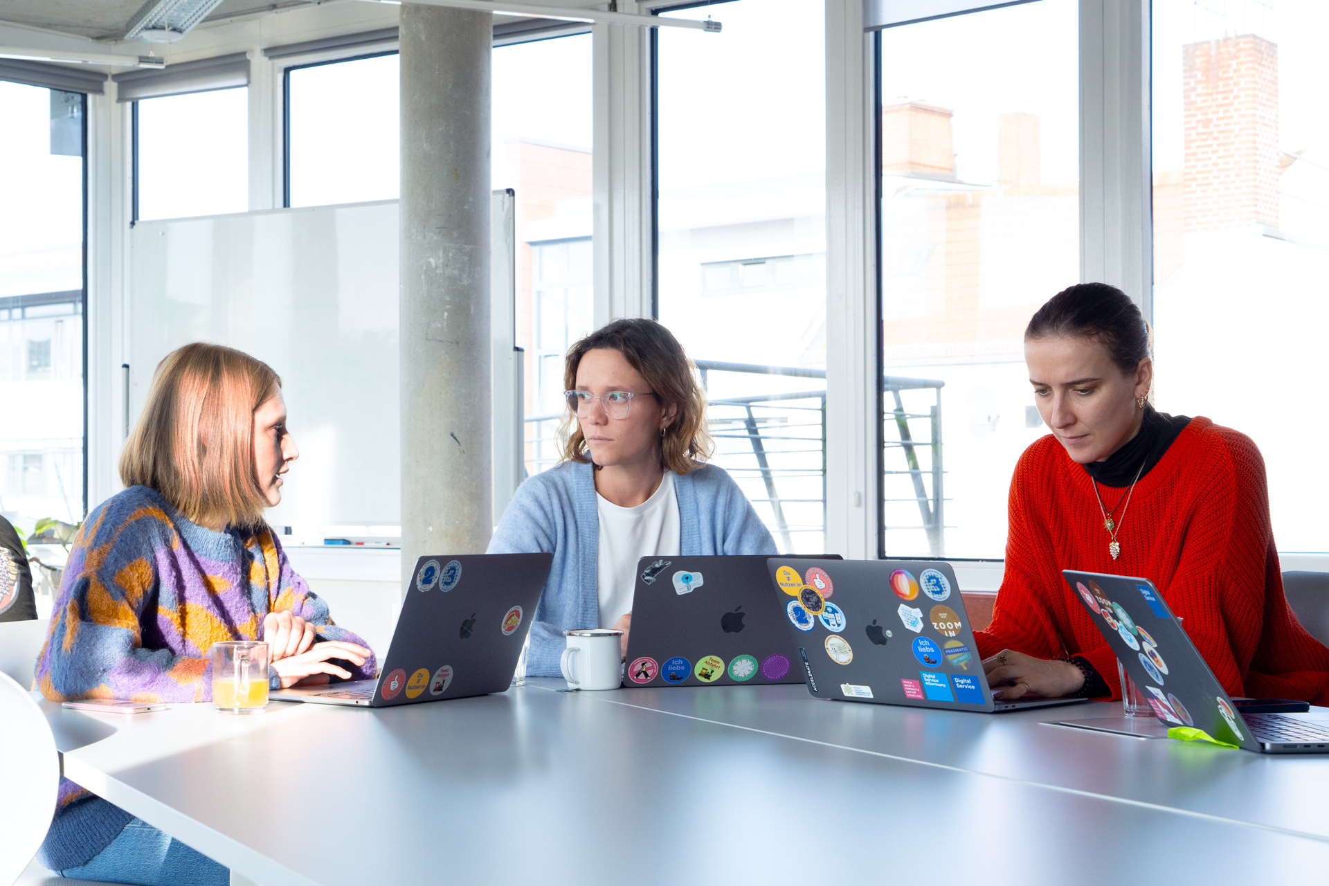 Three DigitalService employees sit together at a large table in the office with their laptops.