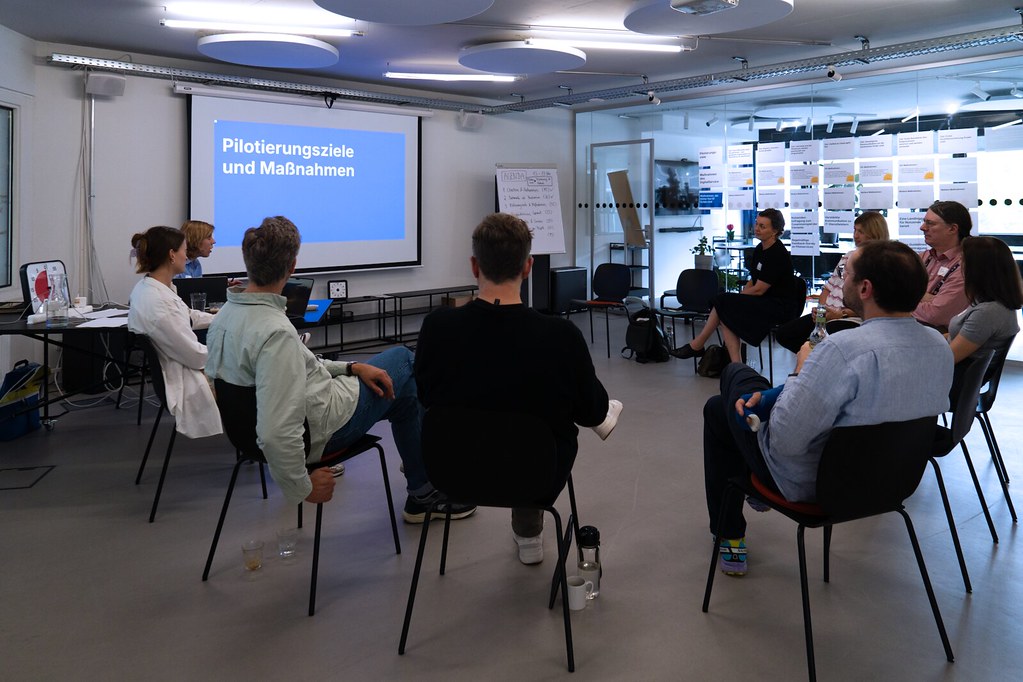 Neun Menschen in einem Stuhlkreis blicken auf eine Präsentation mit der Überschrift „Pilotierungsziele und Maßnahmen“
