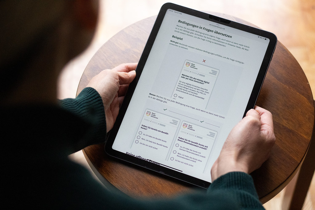 Eine Person hält ein Tablet, das einen Ausschnitt der Servicestandard-Anleitung „Formulare mit verständlichen Fragen gestalten“ anzeigt