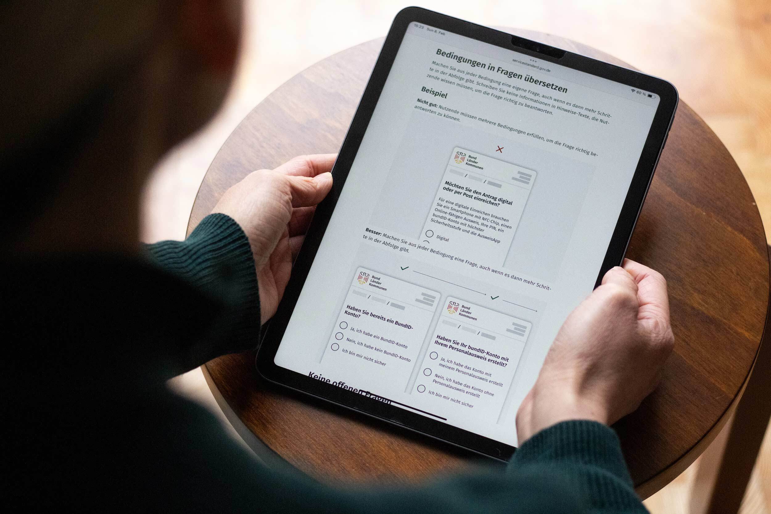 Eine Person hält ein Tablet, das einen Ausschnitt der Servicestandard-Anleitung „Formulare mit verständlichen Fragen gestalten“ anzeigt