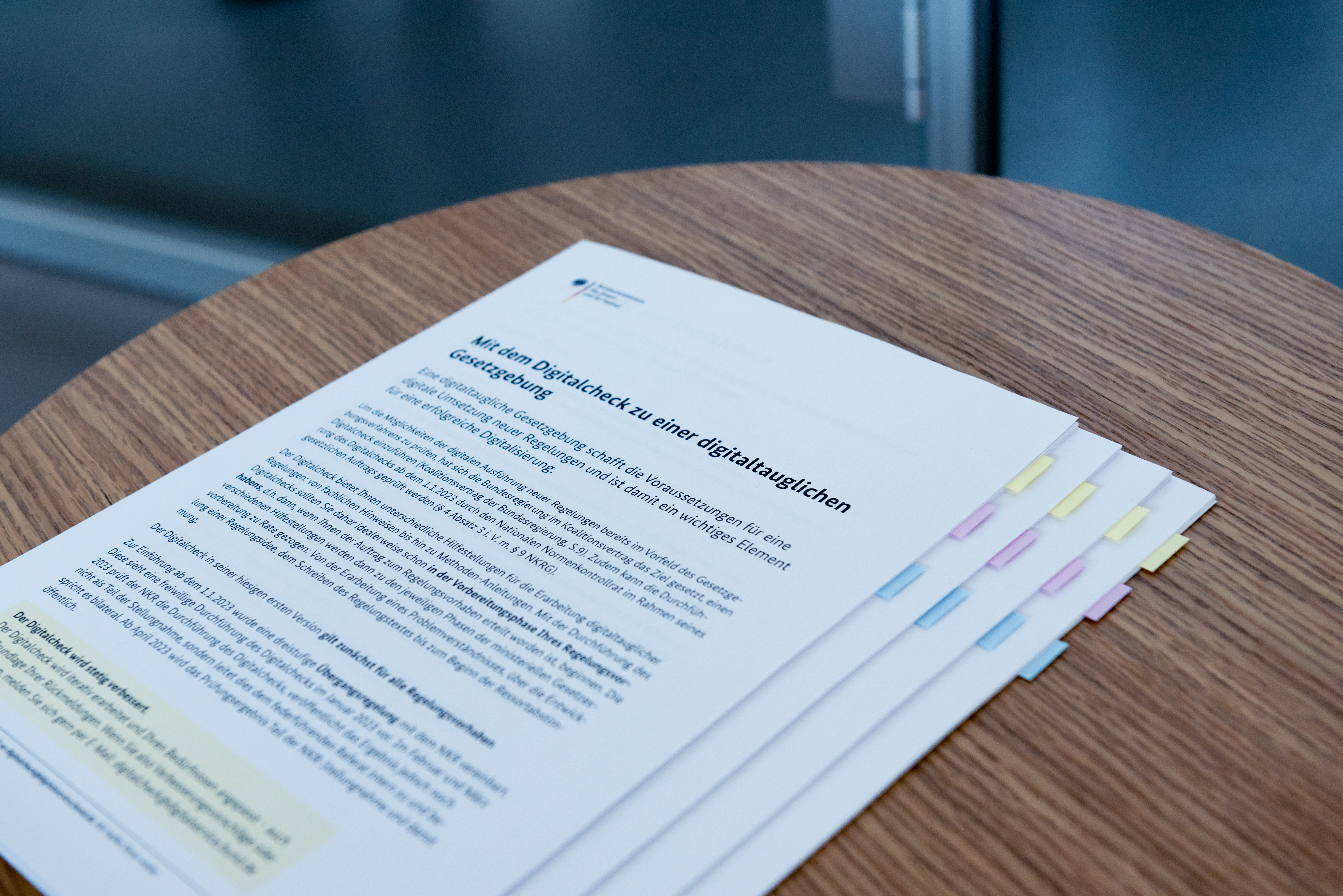 A stack of printed documents with the title “With the digital check to digital-capable legislation” lies on a round wooden table; the pages are marked with colored tabs.