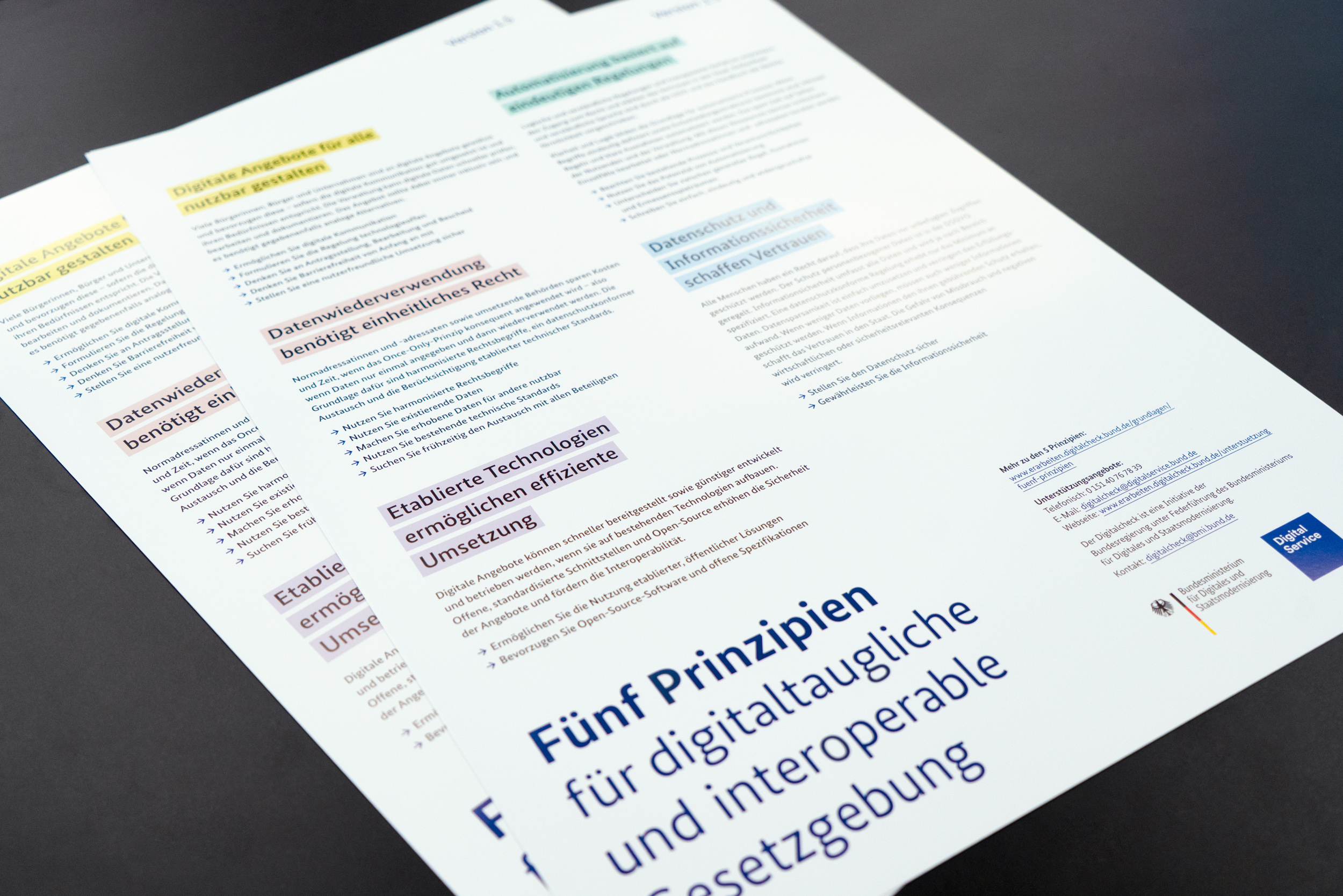 Two posters displaying the five principles for digitally compatible law are laid out on a black table. The text on the posters is highlighted in different colors to distinguish between the various principles. The logos of the Federal Ministry for Digital Transformation and Government Modernisation and DigitalService can be seen in the lower right corner.