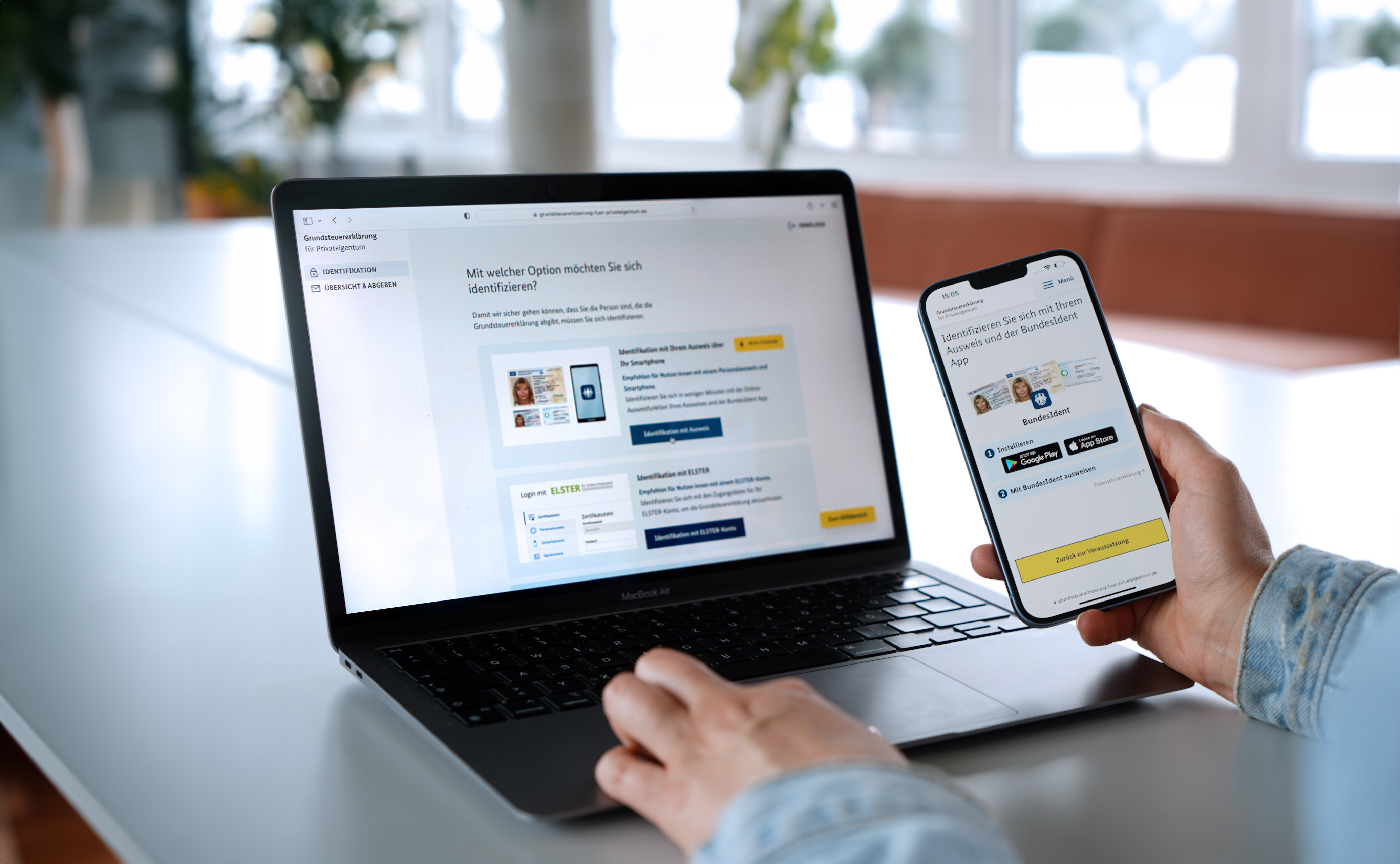 An open laptop displays a website that lists different options for online identification. The person sitting in front of it is also holding a cell phone that displays the BundesIdent start page and is about to use it to identify themself.