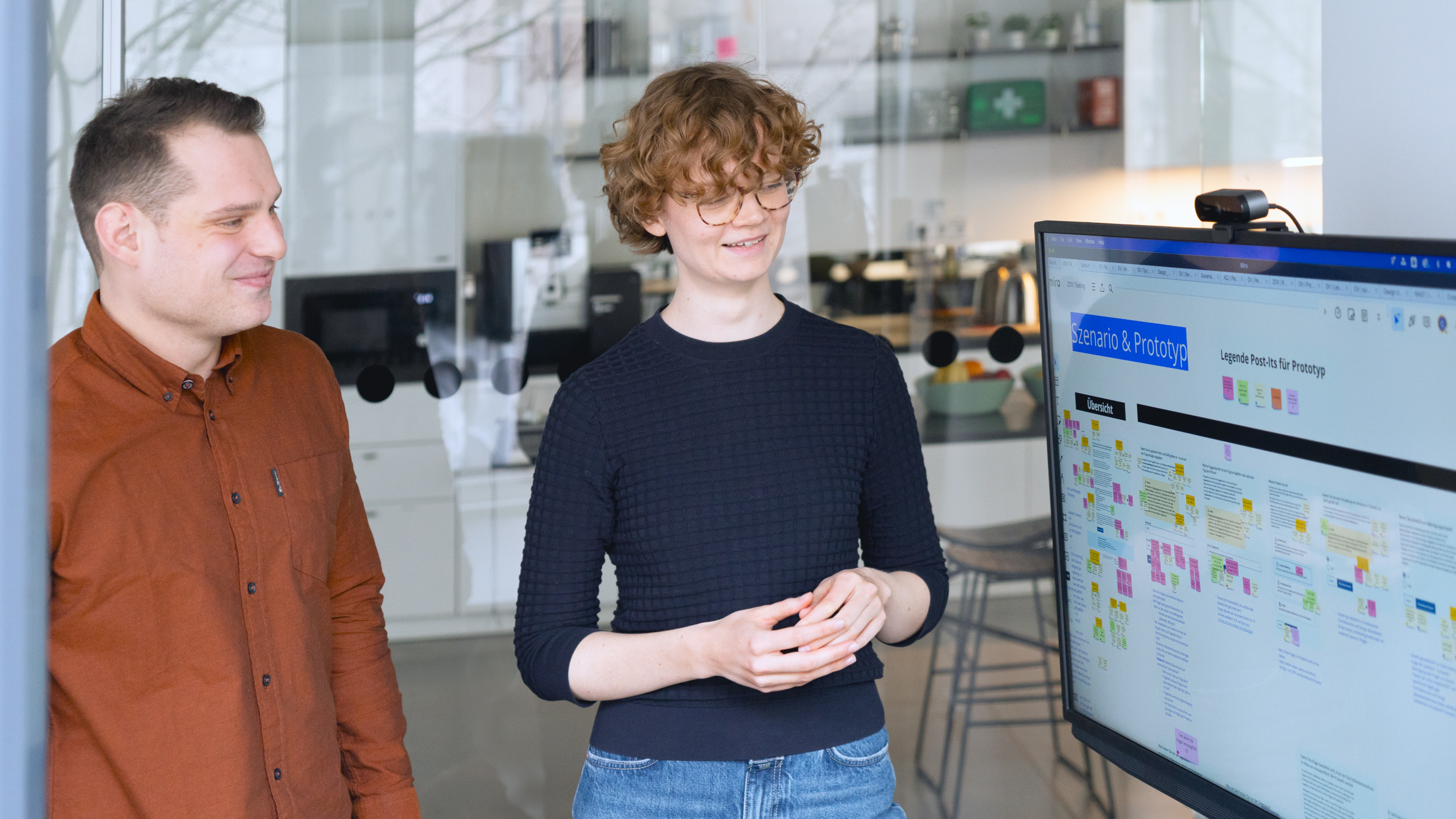 Zwei Personen stehen in einem Meetingraum vor einem großen Bildschirm. Dieser zeigt ein digitales Whiteboard mit der Überschrift „Szenario und Prototyp