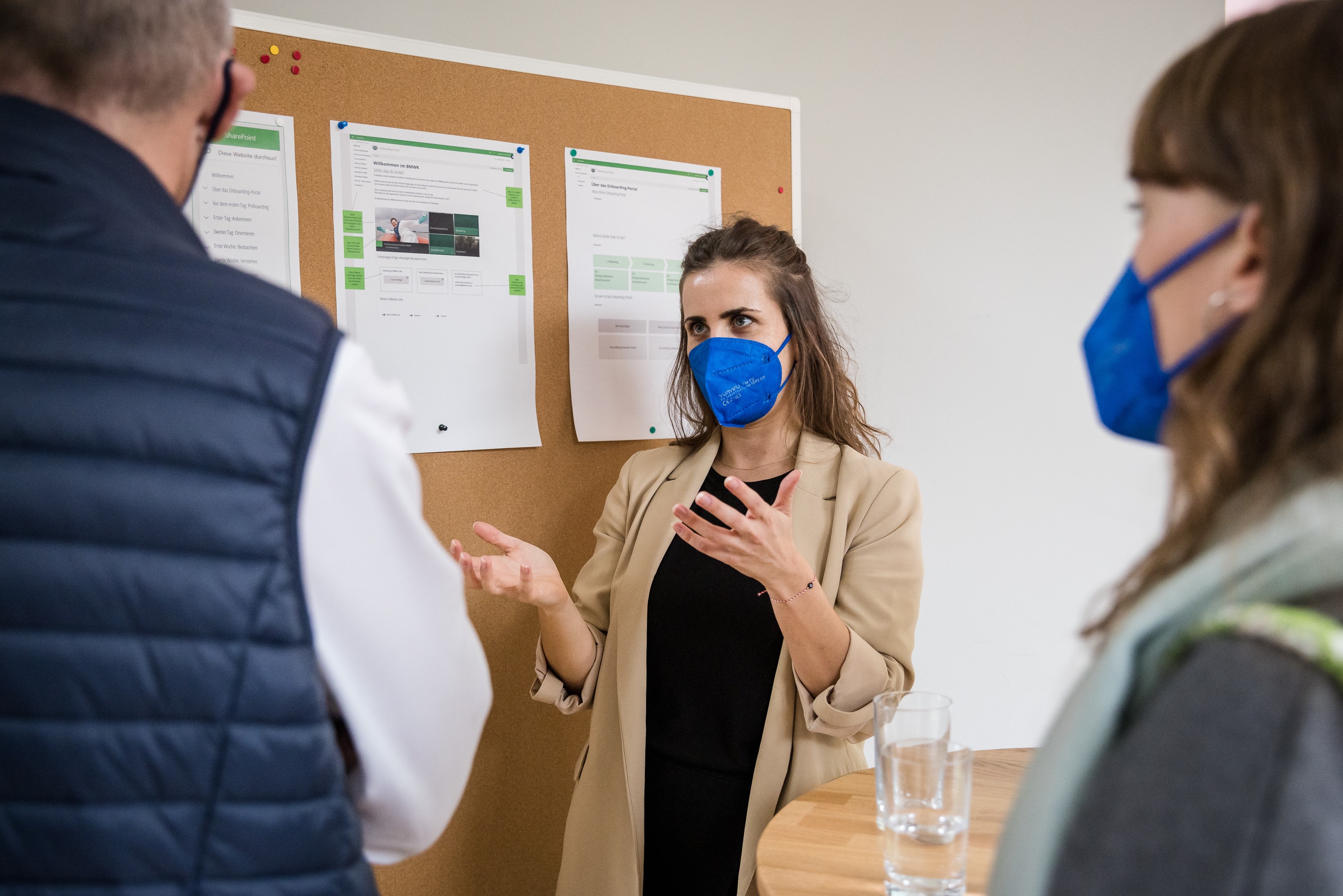 Portraitaufnahme von Lisa. Sie steht vor einer Pinnwand und erklärt zwei Personen ihre Ergebnisse. Dabei trägt sie eine blaue FFP2-Maske.