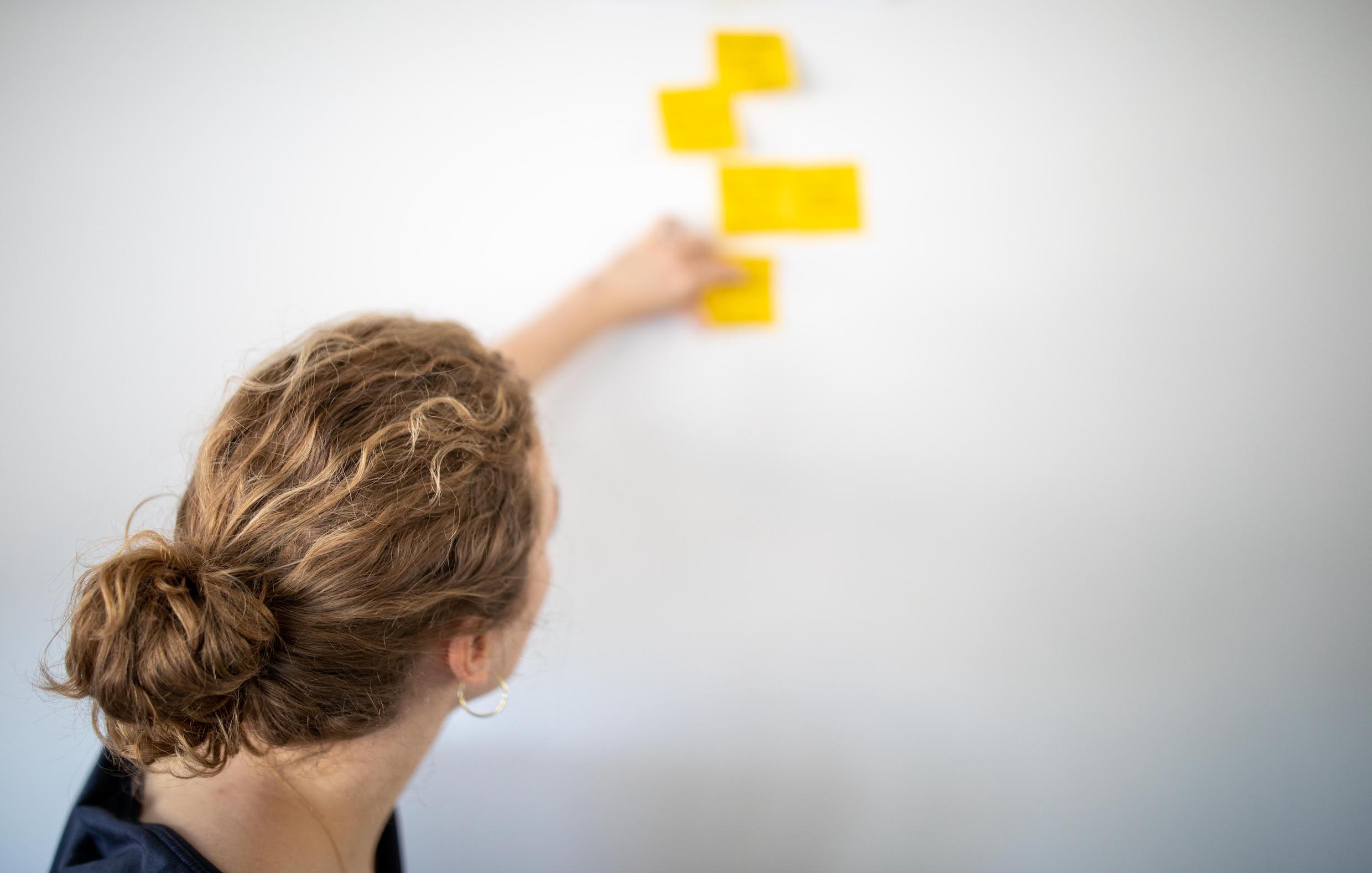 Blonde Frau mit einem Zopf steht nach vorne gerichtet mit dem Geischt zu einem Whiteboard mit gelben Zetteln und befestigt diese am Board.