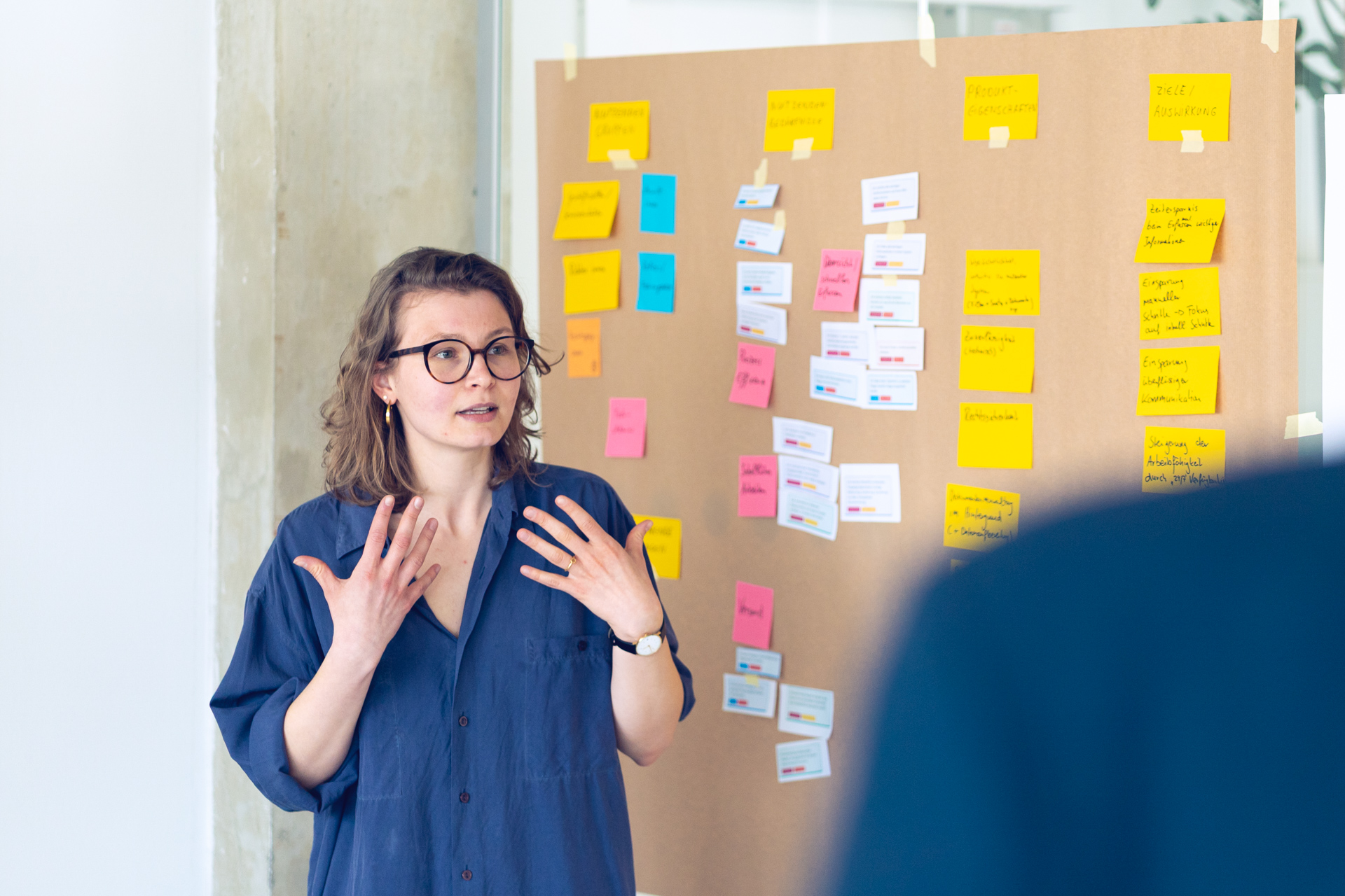 Author Lene Baumgart speaks gesticulatingly in front of a wall covered with colorful sticky notes and cards arranged in columns.