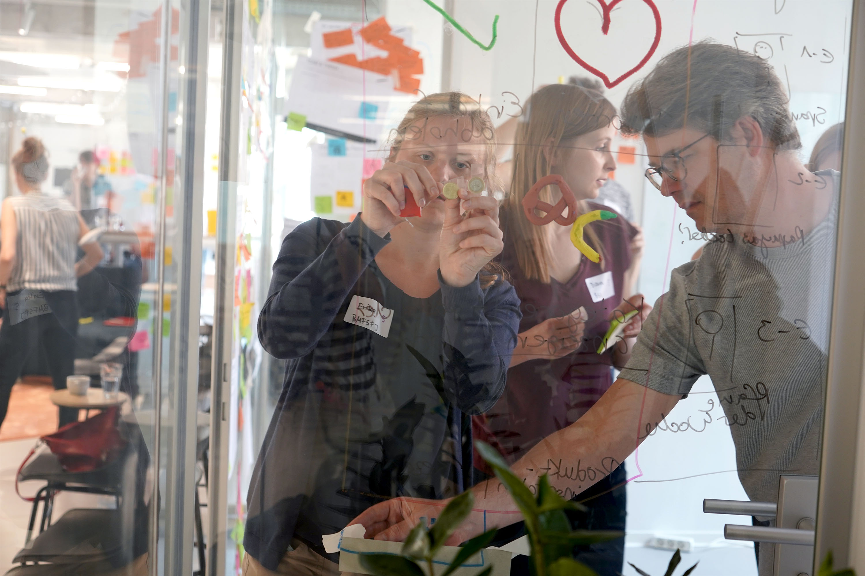 Das Bild zeigt eine Workshop Situation. Das Bild ist durch eine Glaswand durchgeschossen, an die Workshop Teilnehmende schreiben und Dinge drankleben. Die Workshop Teilnehmenden wirken konzentriert bei der Sache.