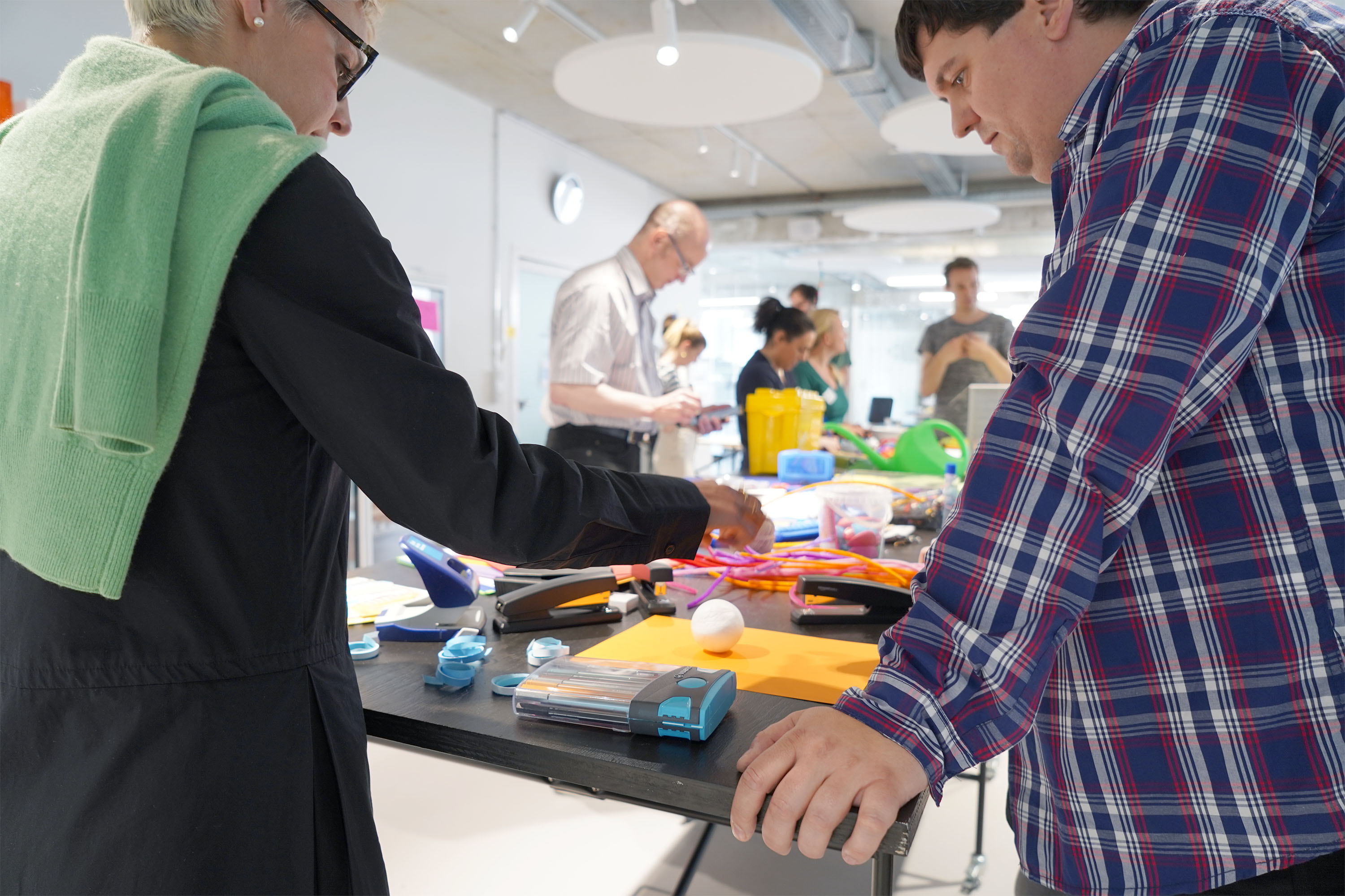 Das Bild zeigt eine Workshop Situation. Teilnehmende stehen um einen großten Stehtisch auf dem Post Its und andere Workshop Materialien liegen. Die Teilnehmenden basteln, probieren aus und beratschlagen sich.