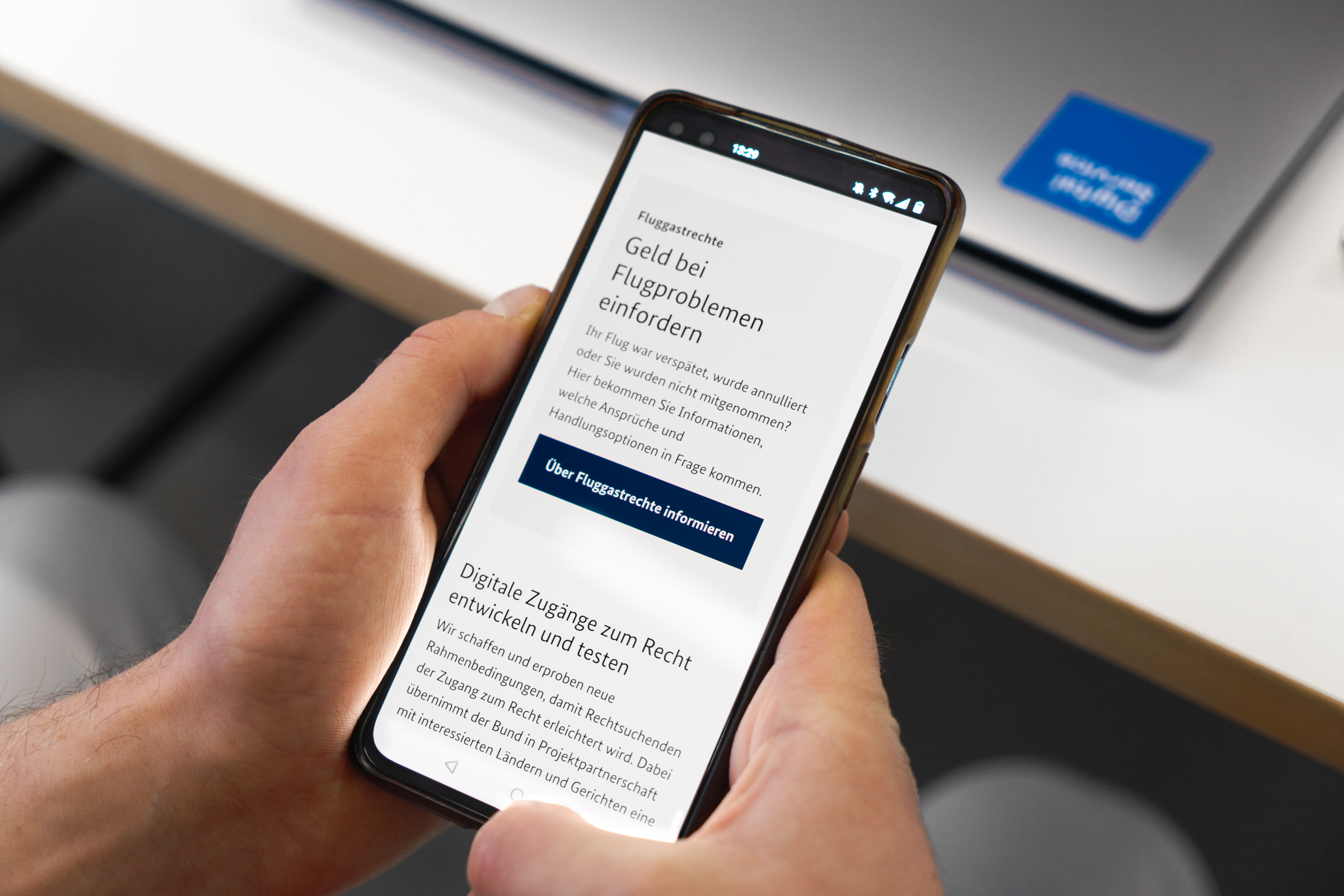 Eine Hand hält ein Smartphone mit einer Website über Fluggastrechte. Der Text zeigt Infos zu Entschädigungen bei Flugproblemen. Im Hintergrund liegt ein Laptop mit blauem Aufkleber auf einem Tisch.