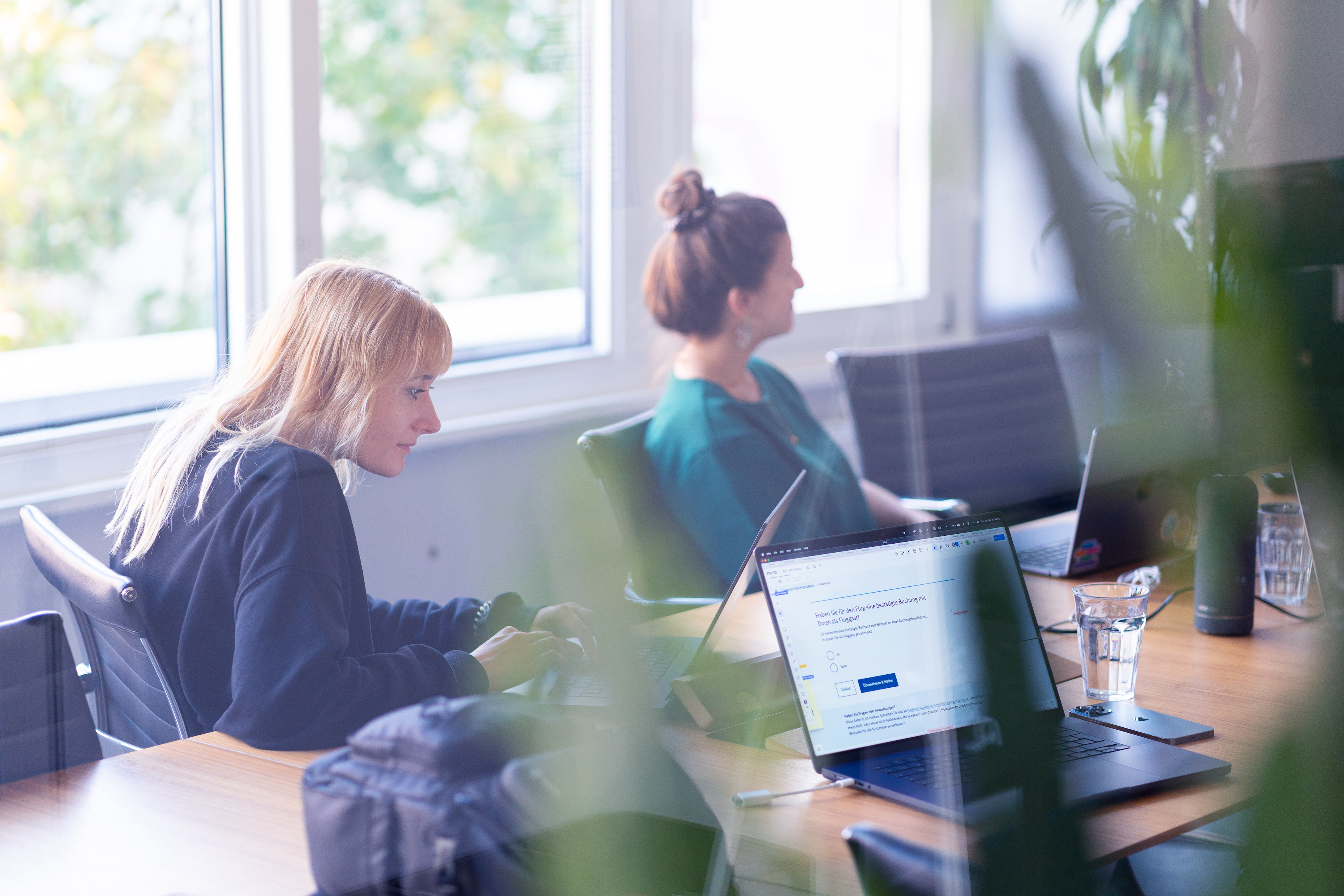 Zwei Personen sitzen an einem Tisch in einem Büro und arbeiten konzentriert an Laptops. Im Vordergrund sind Pflanzen und ein Laptopbildschirm zu sehen, auf dem eine Website geöffnet ist.