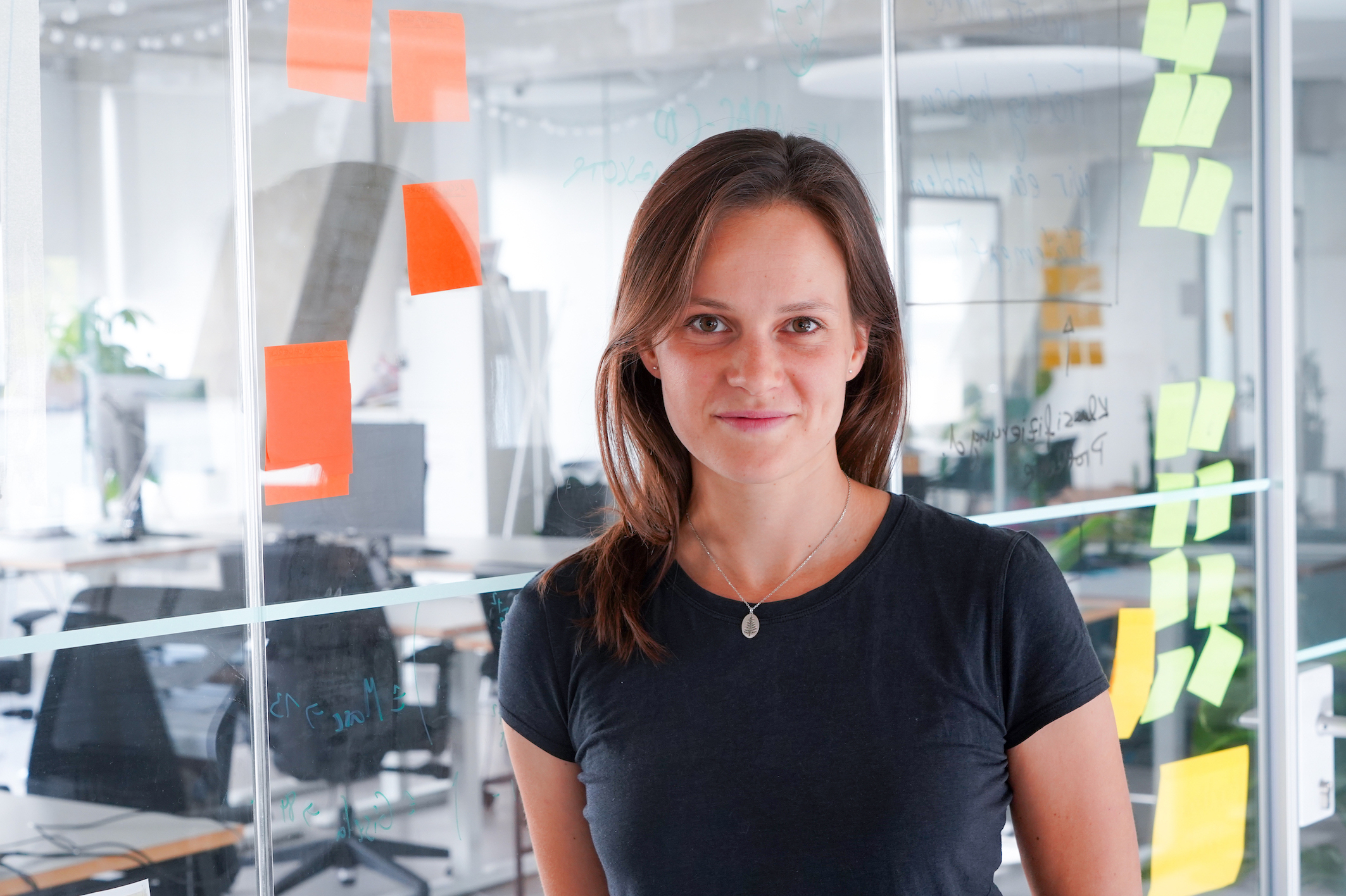 Katja Anokhina, Product Managerin beim DigitalService, steht in einem lichtdurchfluteten Büro vor einer Glaswand; im Hintergrund sind Tische und Bürostühle zu sehen; sie trägt ein schwarzes T-Shirt und hate braune schulterlange Haare