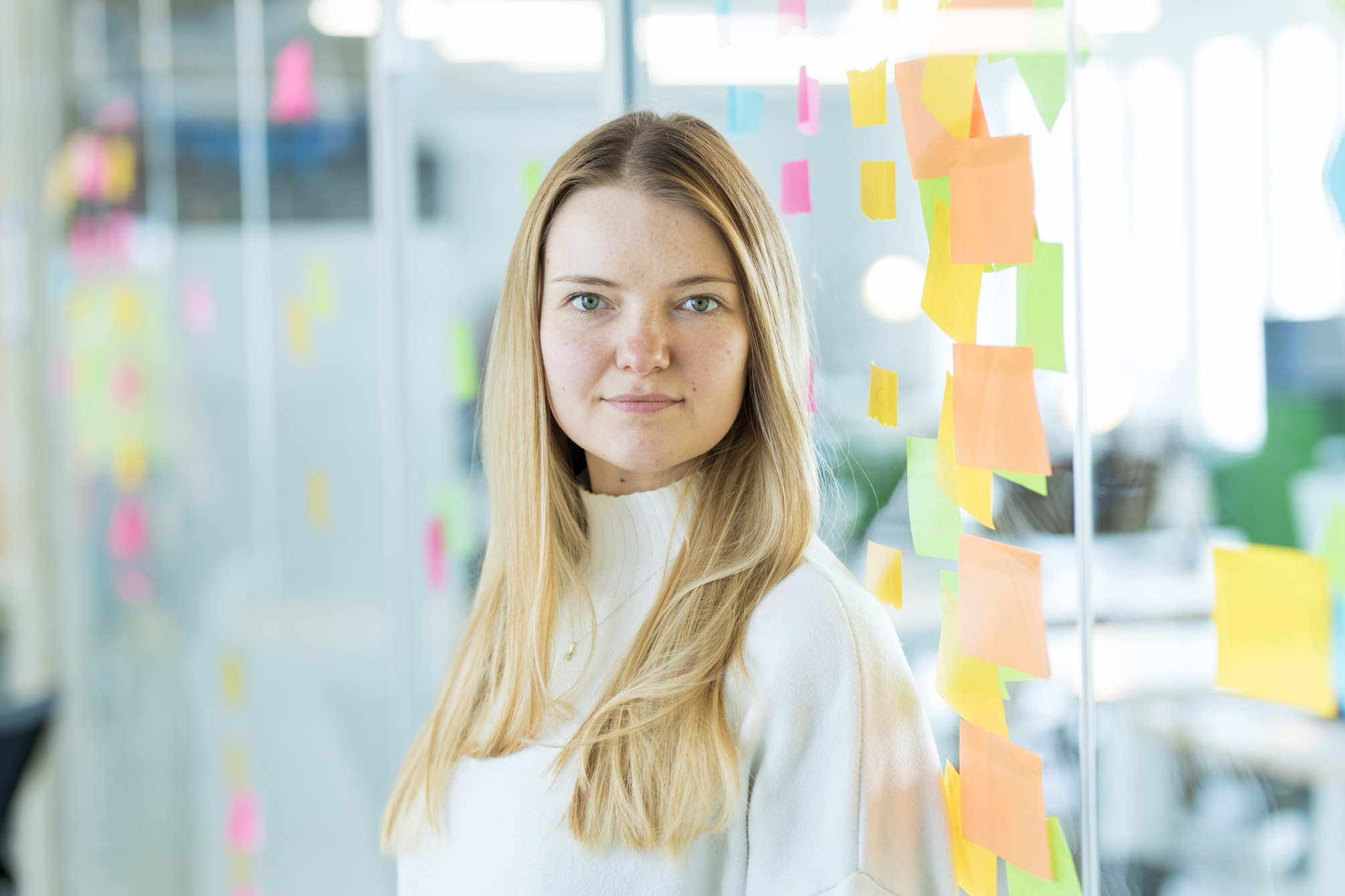 Carolin, Produktmanagerin beim DigitalService, steht im Büro des Unternehmens vor einer Glaswand, die mit vielen bunten Notizzetteln verziert ist. Sie hat blonde, lange Haare und trägt einen hellen Rollkragenpullover.