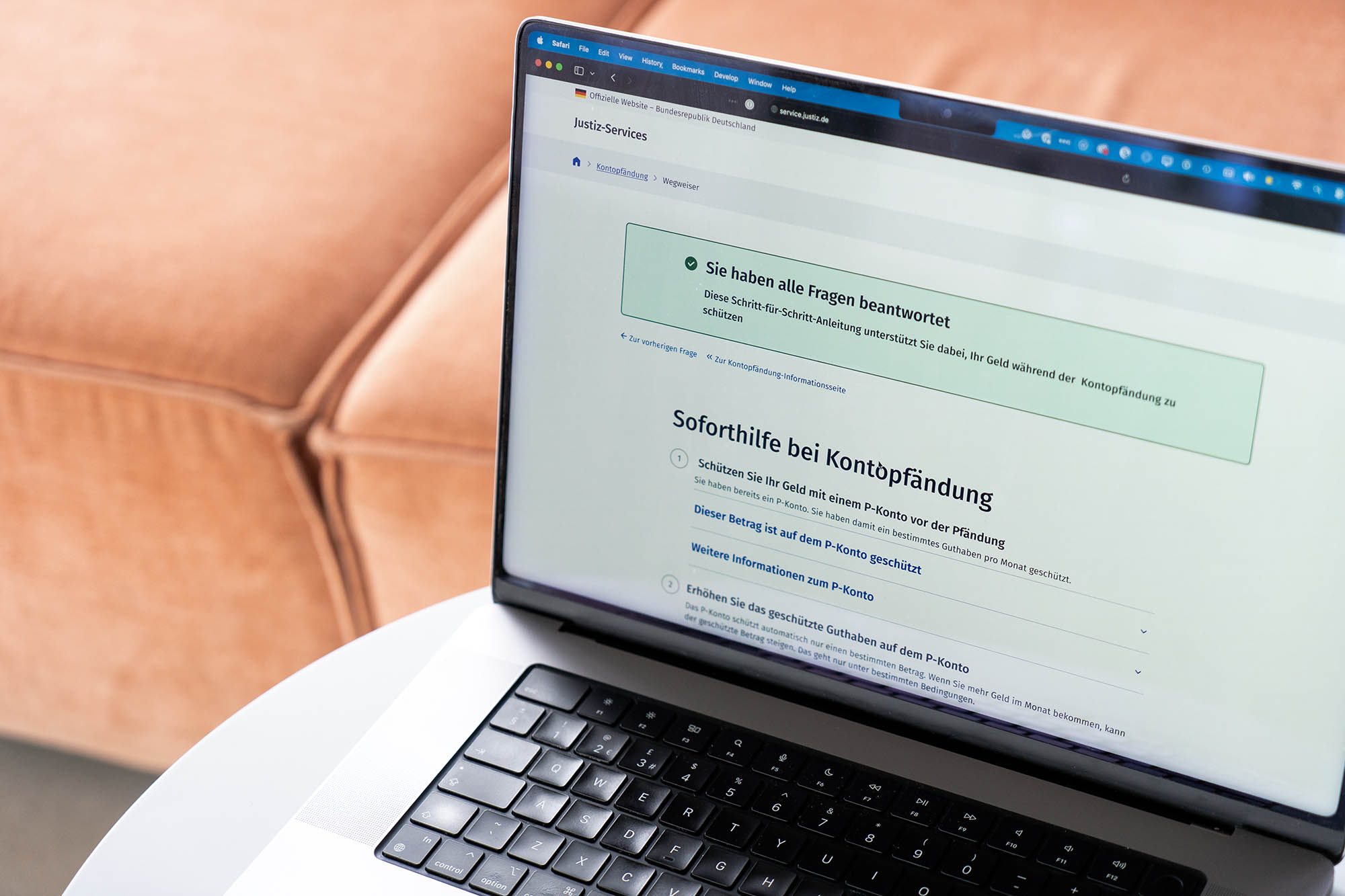 Laptop auf einem kleinen Tisch, es zeigt eine Bestätigung eines Justiz-Services, der sagt: ,Sie haben alle Fragen beantwortet – Diese Schritt-für-Schritt-Anleitung unterstützt sie dabei, Ihr Geld während der Kontopfändung zu schützen‘, darunter: Sorforthilfe bei Kontopfändung mit einer nummerierten Liste