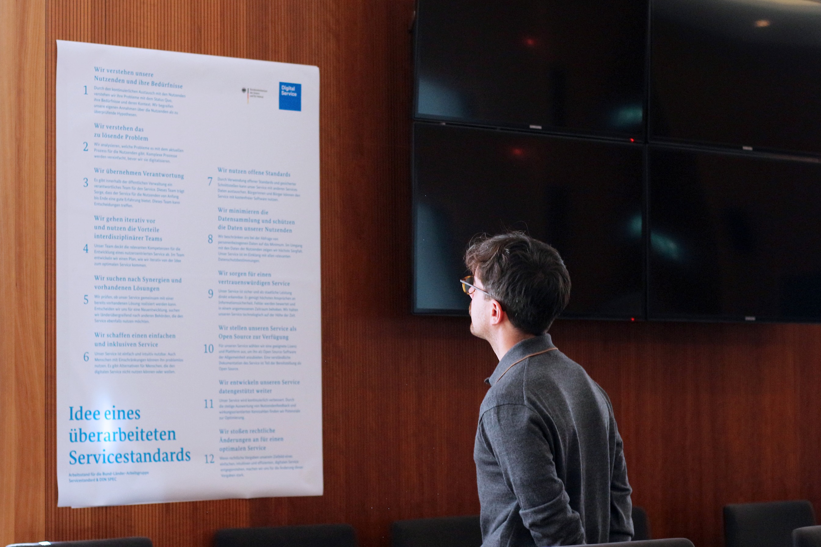 A person looks at a large, white poster on a wooden wall. The poster shows various points of the idea of a revised service standard with a blue heading and text. Black screens hang next to it.