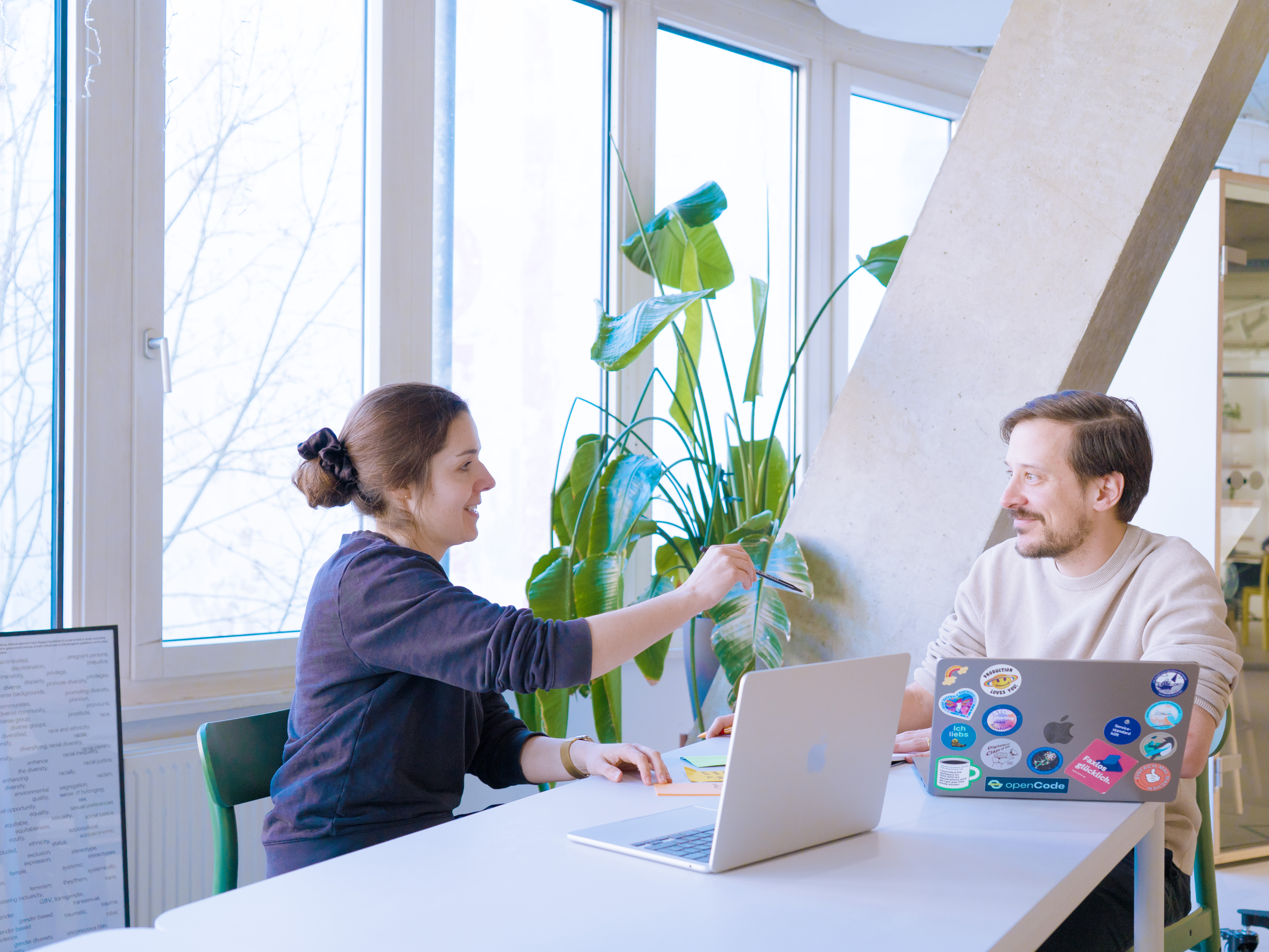 Foto von Magdalena Zadara und Niko Felger in einem modernen Büroraum. Beide sprechen miteinander, Magdalena gestikuliert mit einem Stift. Vor den beiden stehen auf einem Tisch Laptops mit geöffneten Bildschirmen.