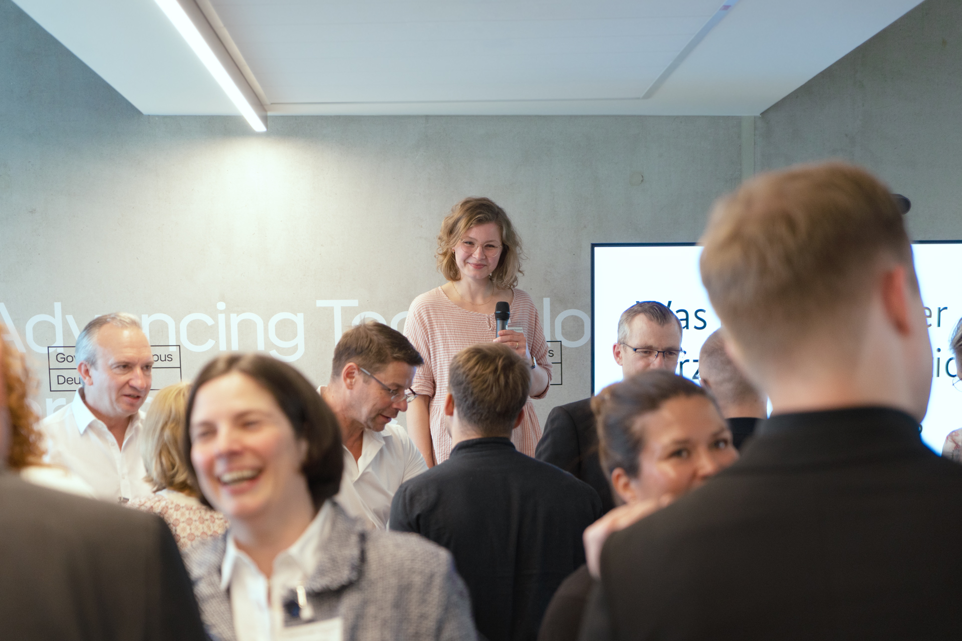 Lene Baumgart steht mit einem Mikrofon vor einer Gruppe von Menschen, die sich in einem modernen Konferenzraum unterhalten. Im Hintergrund ist ein Bildschirm mit Text und eine Wand mit Schriftzügen zu sehen.