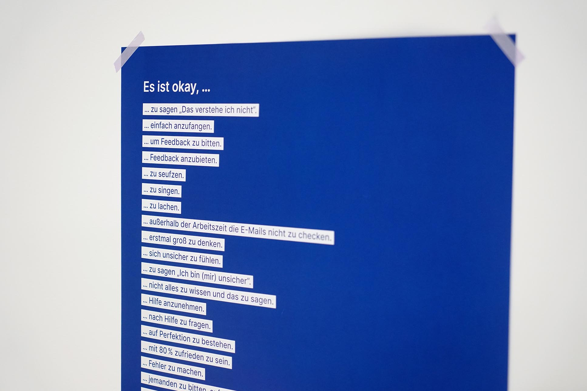 Ein blaues Plakat mit lila Klebestreifen an einer Wand angebracht, überschrieben mit: Es ist okay, Punkt, Punkt, Punkt und einer Auflistung von Dingen, wie: zu sagen, „Das verstehe ich nicht”, „einfach anzufangen“, “um Feedback zu bitten”