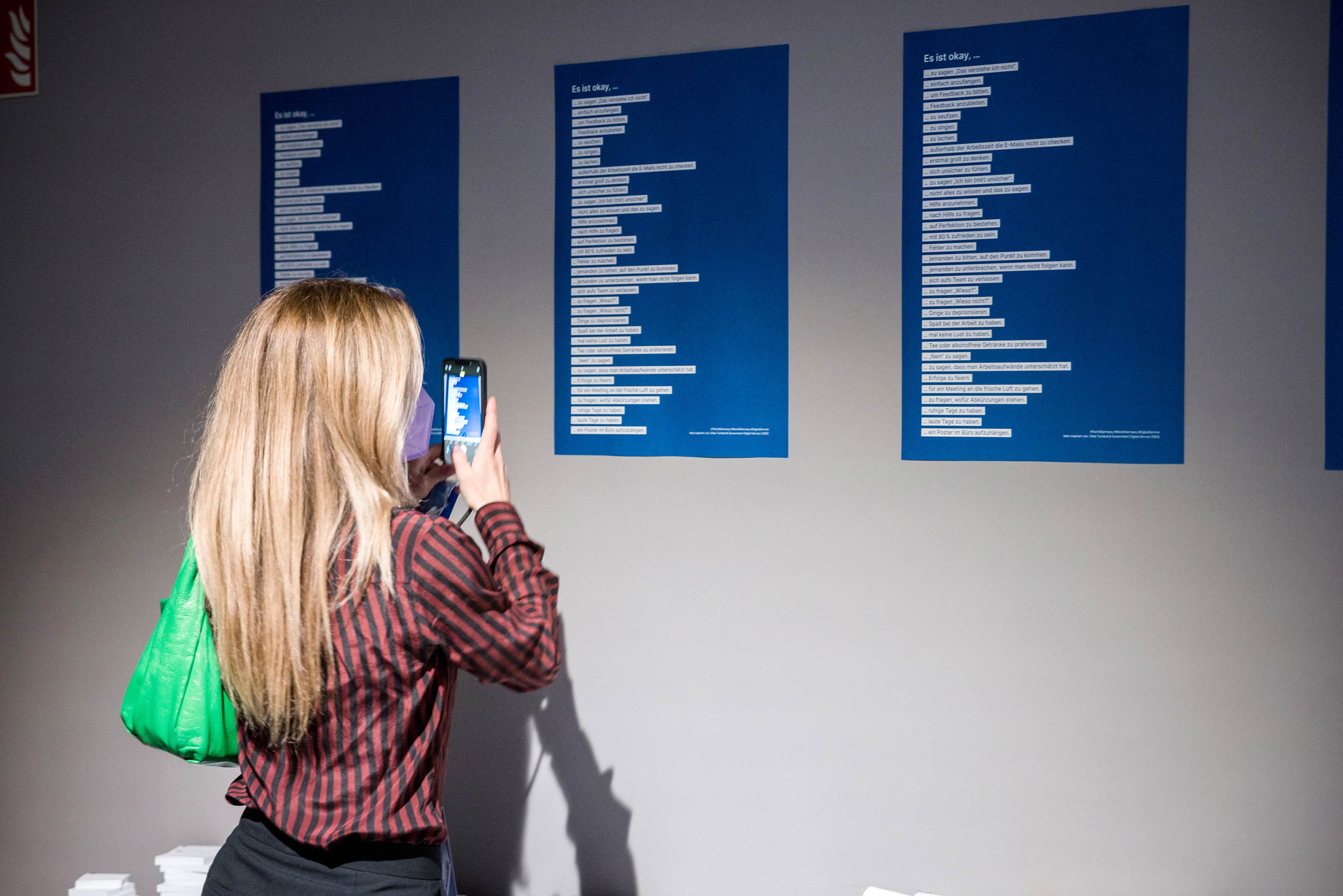 A person with long light hair stands in front of three identical blue posters, which she photographs with her phone - the posters contain two dozen lines of text