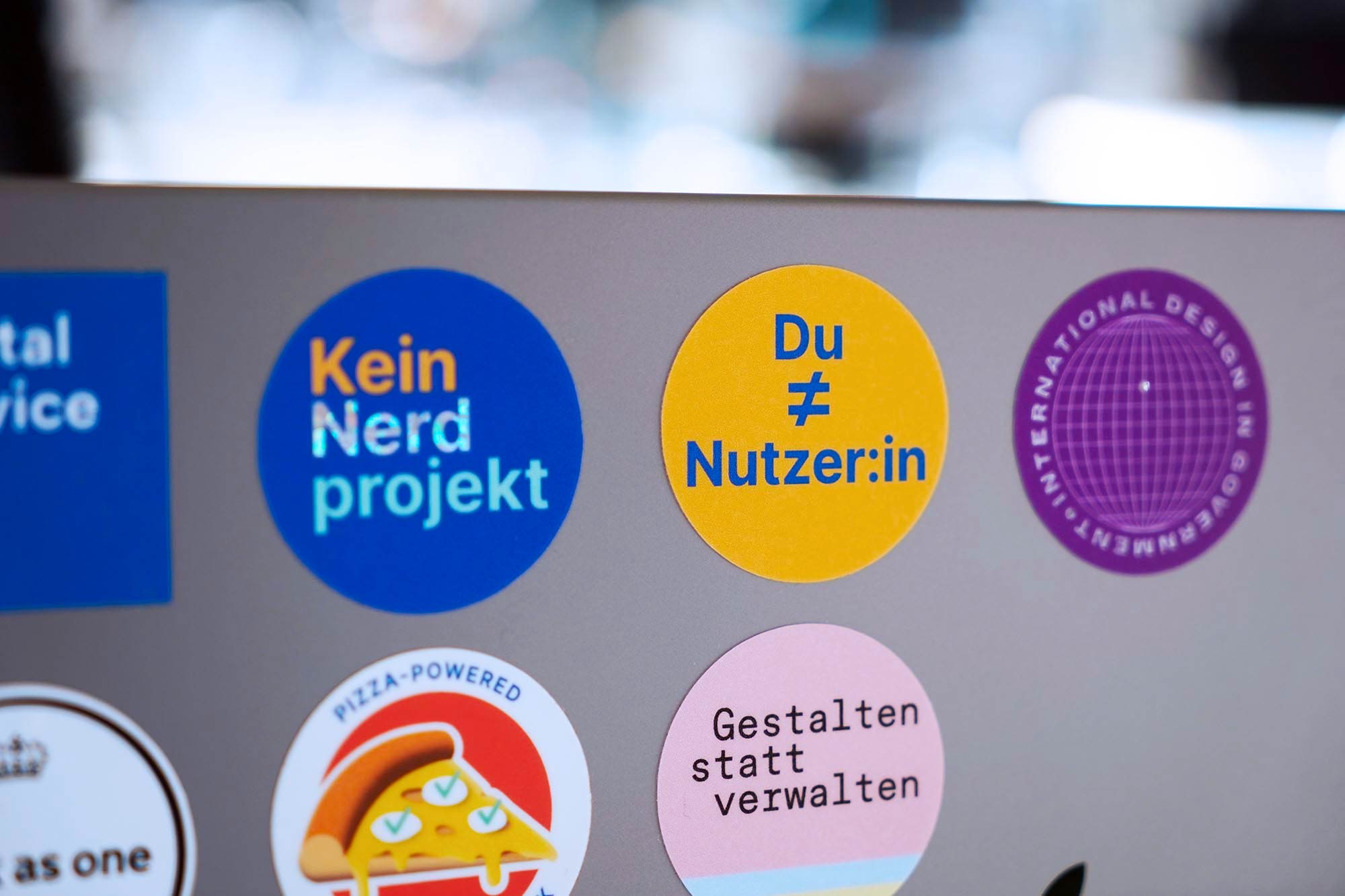 Mehrere farbige Sticker auf einem grauen Laptop-Computer – die Sticker im Fokus sagen ,Kein Nerdprojekt‘, ,Du ungleich Nutzer:in‘ und ,Gestalten statt verwalten
