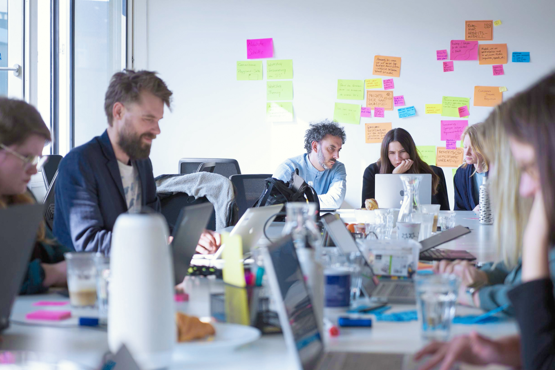 7 Personen mittleren Alters sitzen mit ihren Laptops an einem langen Tisch, einige unterhalten sich miteinander, andere schauen auf ihre Computer, an der Wand hinter ihnen kleben verschiedene bunte Haftnotizen.