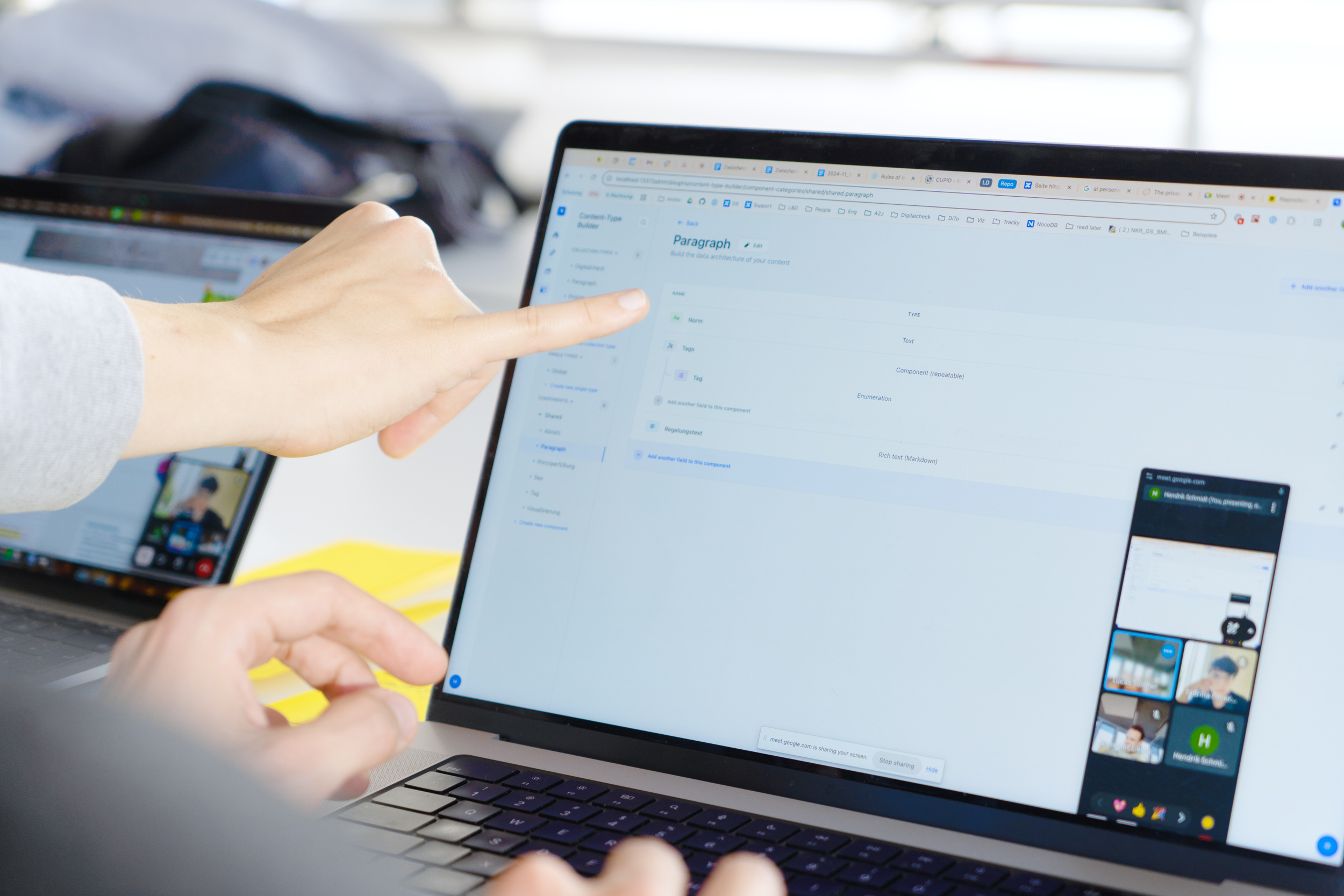 Eine Person zeigt mit dem Finger auf den Bildschirm eines Laptops, auf dem eine Benutzeroberfläche mit dem Titel „Paragraph“ zu sehen ist, während im Hintergrund ein zweiter Laptop mit geöffnetem Videoanruf sichtbar ist.