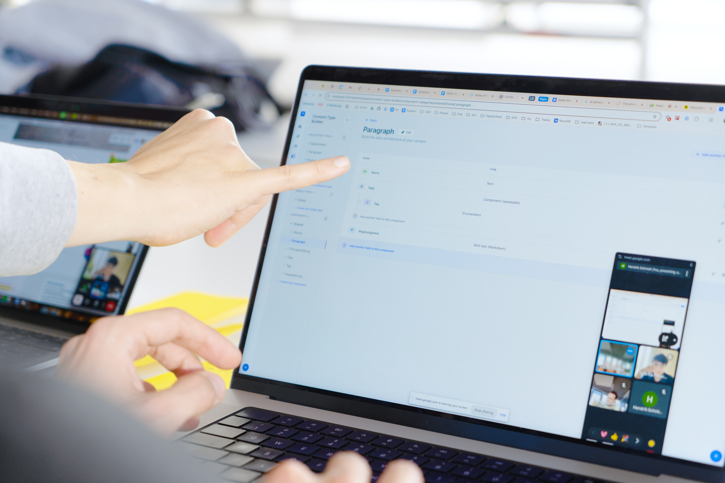 A person points a finger at the screen of a laptop on which a user interface with the title “Paragraph” can be seen, while a second laptop with an open video call is visible in the background.