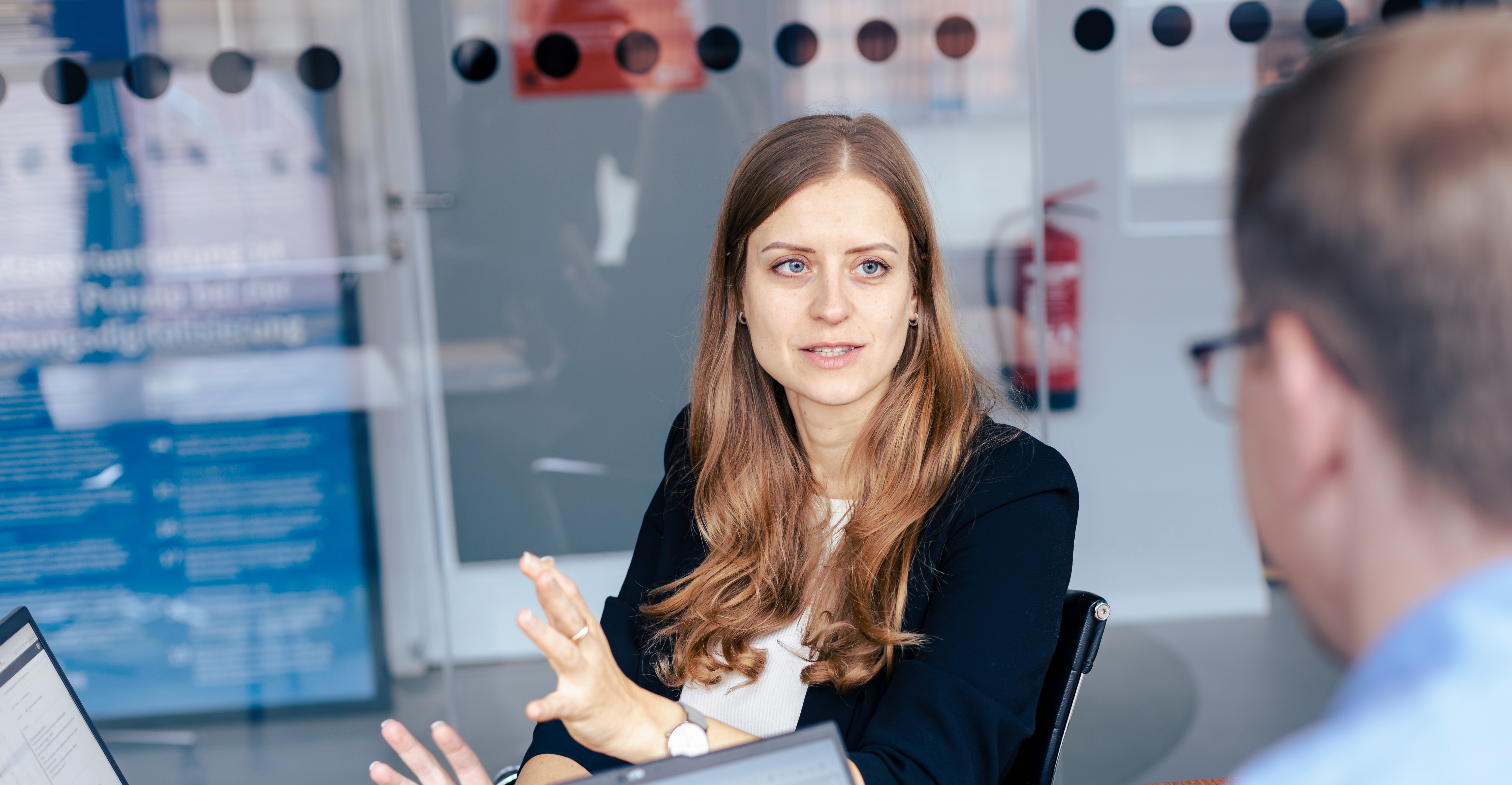 Christina Lang spricht in einem modernen Büro mit Glaswänden mit einem Kollegen. Sie trägt einen schwarzen Blazer und eine helle Bluse und gestikuliert.