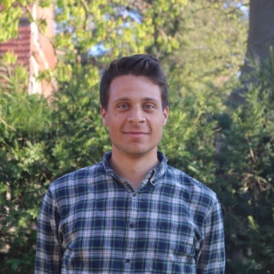 Sami Yacob, Product Manager beim DigitalService, trägt ein blau kariertes Hemd und steht in der Natur vor einem Baum mit grünen Blättern; er hat dunkle kurze Haare