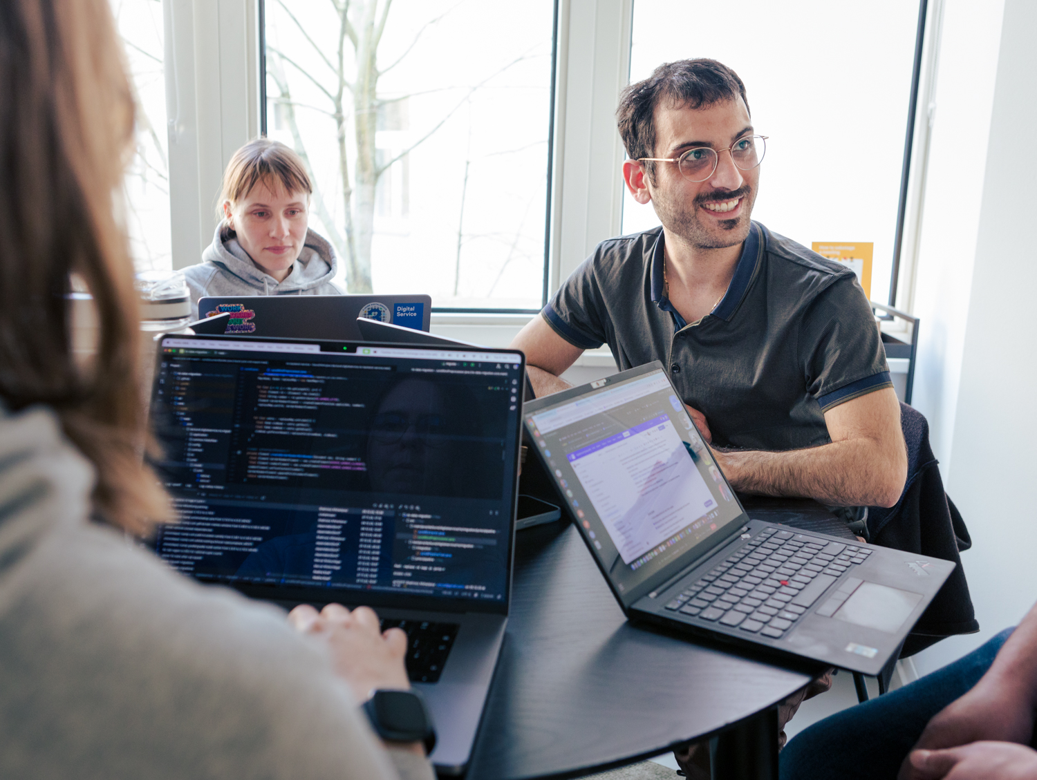 Four people are sitting around a table with their laptops. Lines of code can be seen on the front screen. A man sits in the background and smiles to the right at a person at the table whose face is not visible. To the left of the man, a woman is looking at her screen.