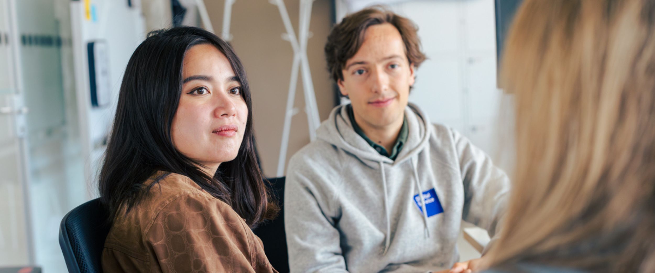 Two DigitalService employees are sitting in an office looking at a third person who can only be seen from behind. In the background are a coat rack, cabinets, and a bulletin board.