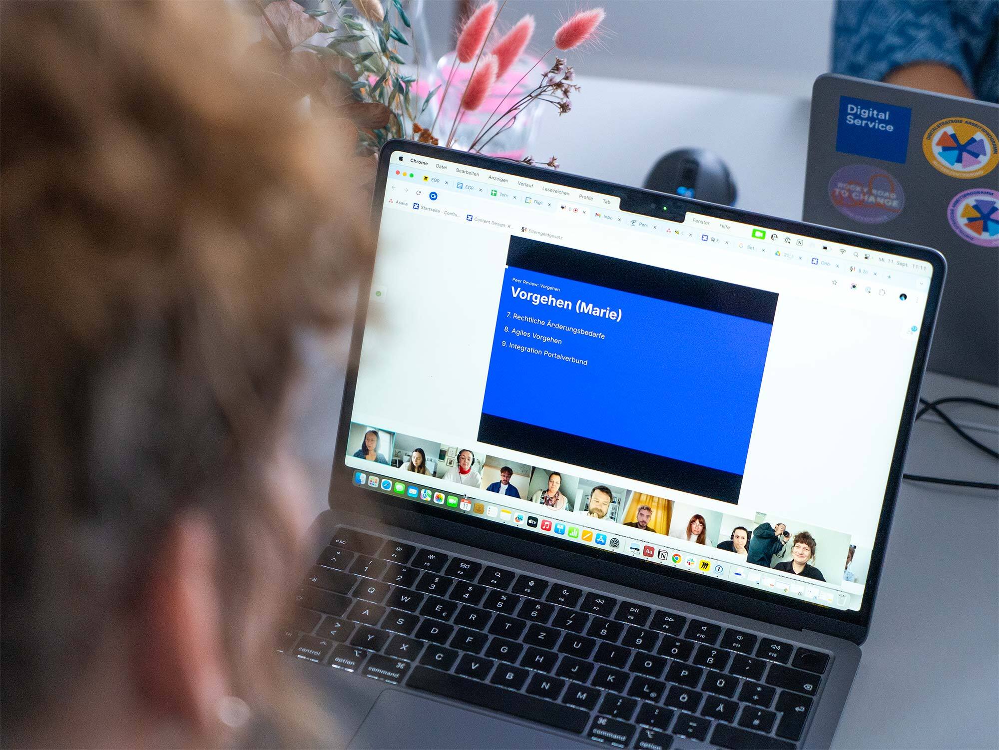 Eine Person nimmt an einer virtuellen Besprechung auf einem Laptop teil. Auf dem Bildschirm wird eine Präsentationsfolie mit dem Titel „Vorgehen (Marie)“ und einer Liste von Aufzählungspunkten angezeigt. Unter der Folie sind die Miniaturansichten der Videos mehrerer Teilnehmer in einer Reihe zu sehen. Im Hintergrund arbeitet eine andere Person an einem Laptop.