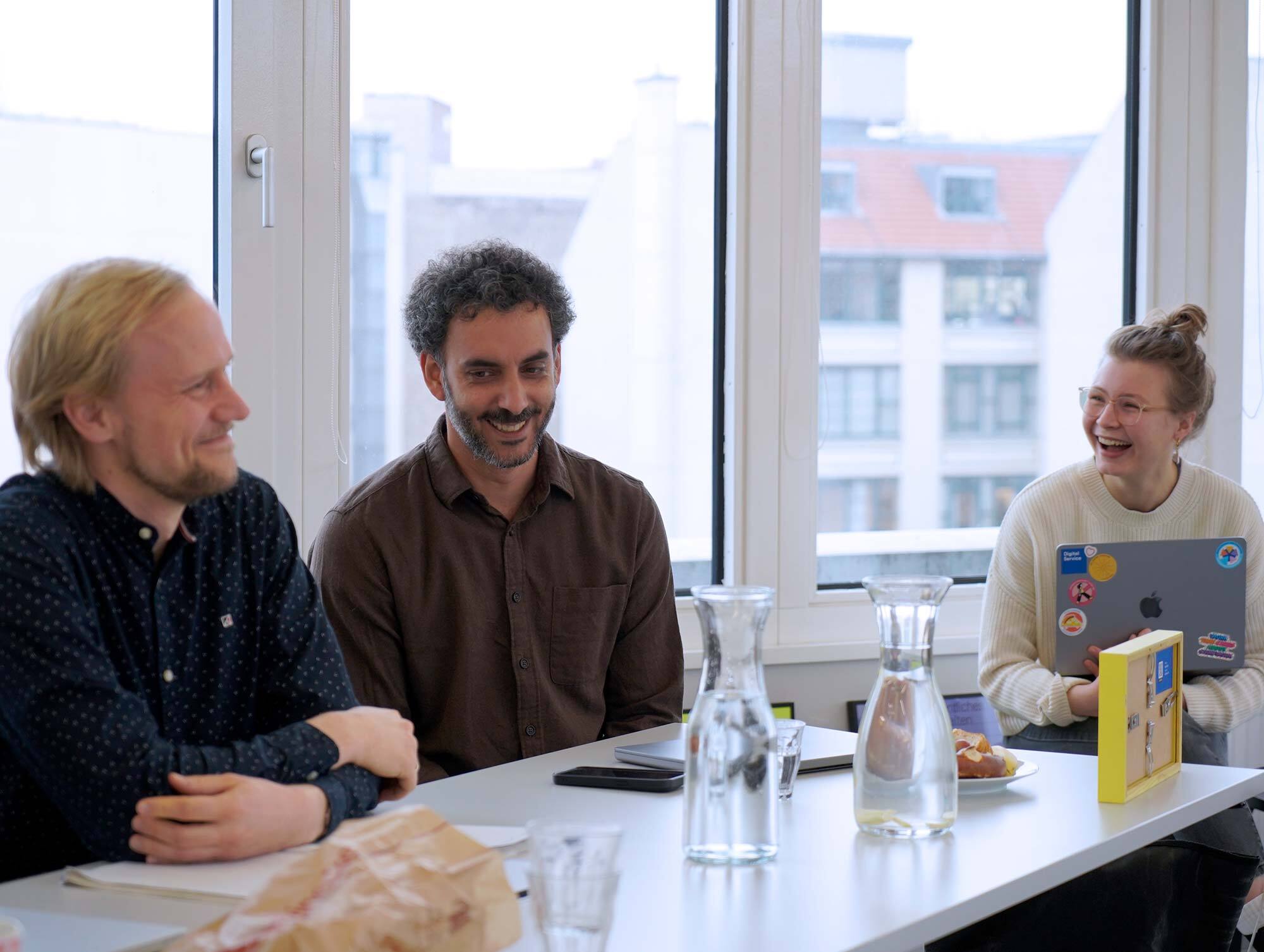 Drei Personen sitzen lächelnd an einem Tisch in einem modernen Büro mit großen Fenstern. Auf dem Tisch stehen Karaffen mit Wasser, Notizbücher und ein Laptop mit Aufklebern.