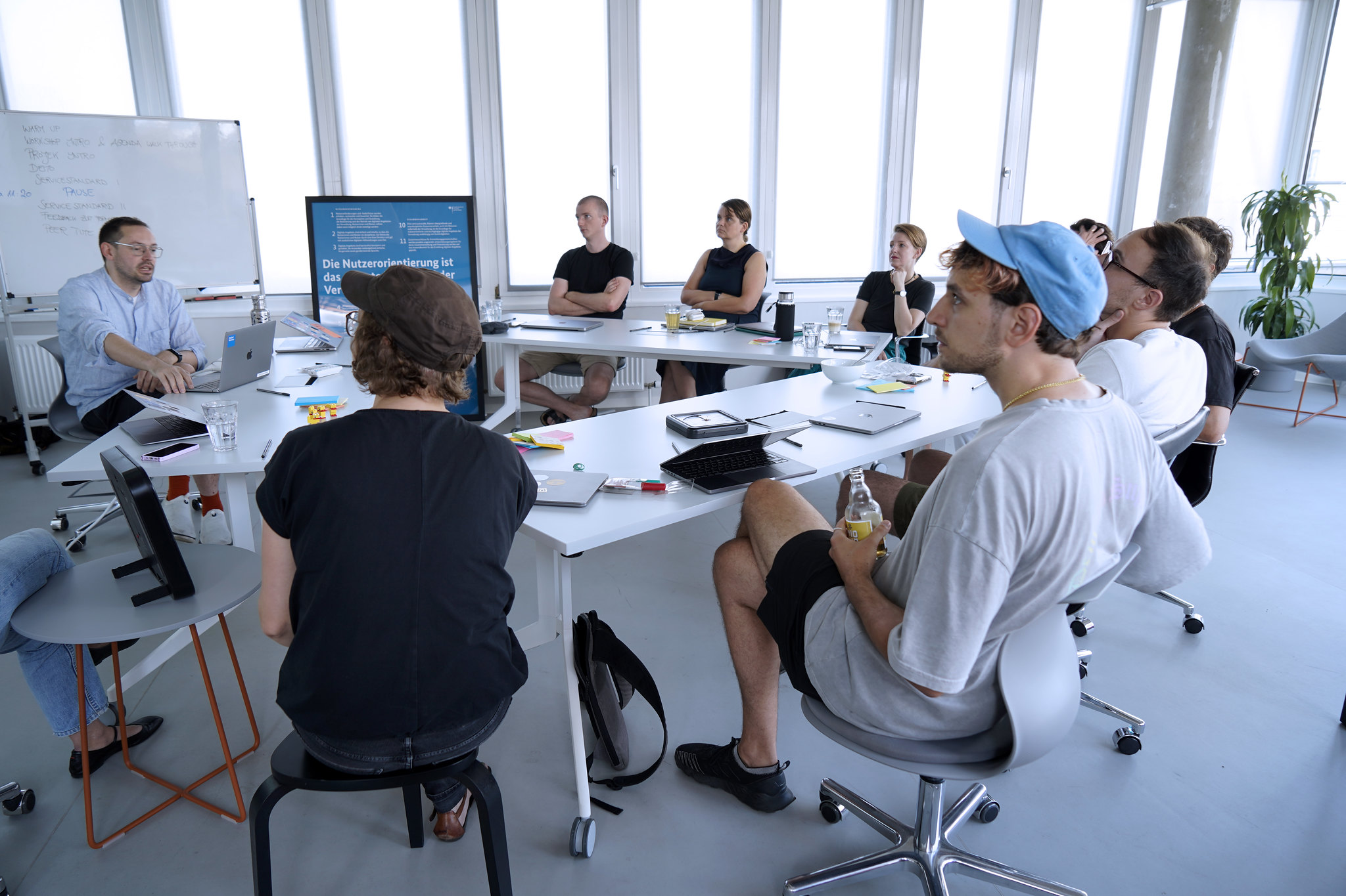 Eine Gruppe von 8 Personen in informeller Kleidung, die in einem modernen, lichtdurchfluteten Büro um zu einem Dreieck aufgestellten Tisch herum sitzen mit einer Projektorfläche im Hintergrund