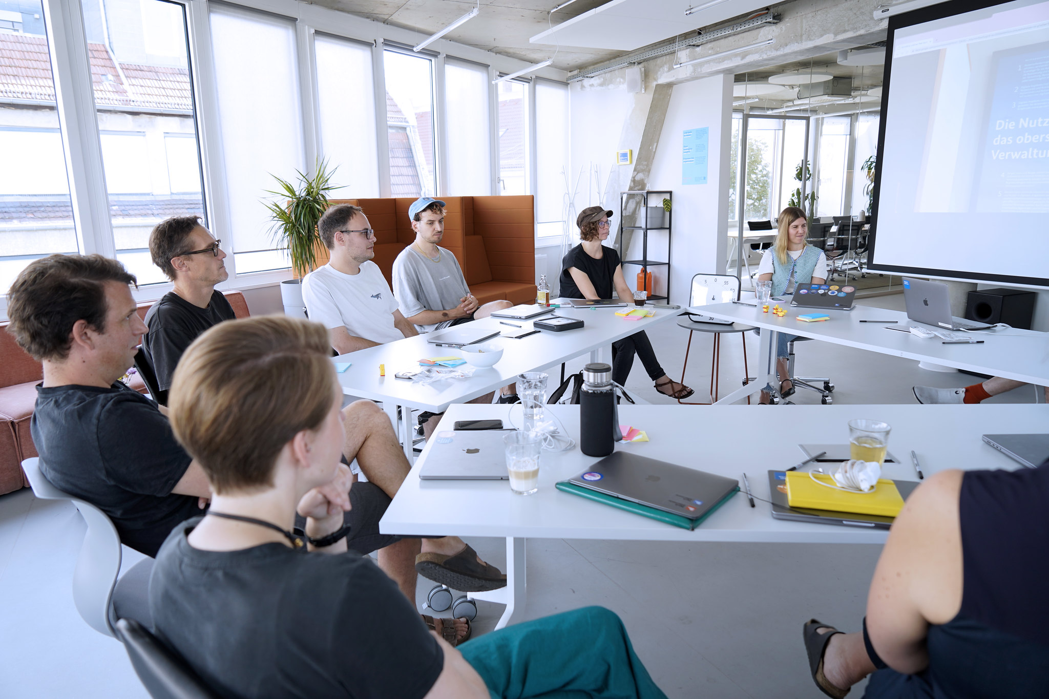 Eine Gruppe von 10 Personen in informeller Kleidung, die in einem modernen, lichtdurchfluteten Büro um einen Tisch herum sitzen – mit einem Whiteboard und einem Poster des Servicestandards hinter ihnen